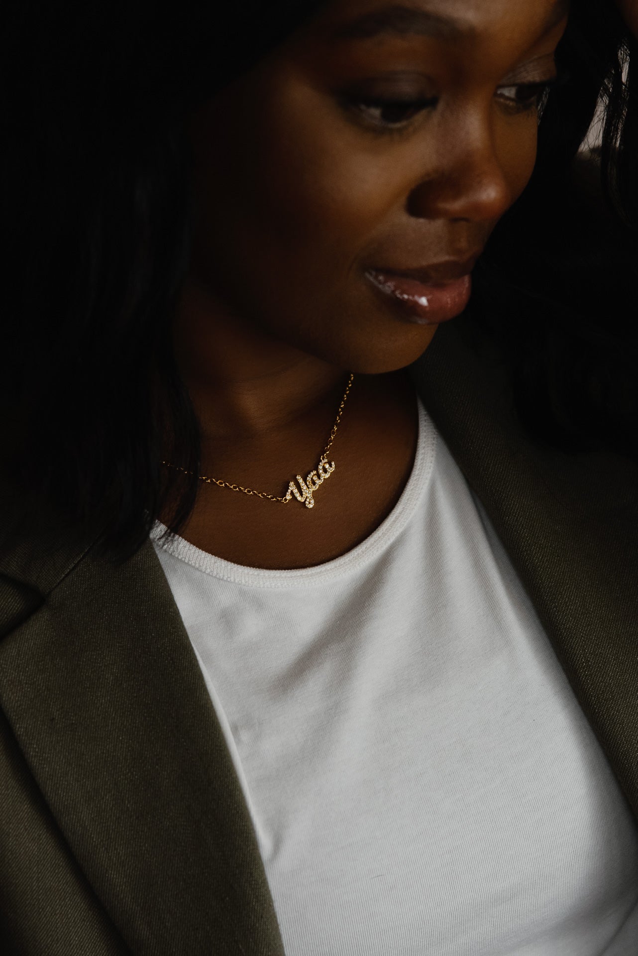 A close-up of a model wearing CASA London's 18k gold Yaa Pavé Necklace. The delicate gold chain features a pendant with the name "Yaa" intricately encrusted with shimmering pavé-set stones, celebrating the tradition of those born on a Thursday. The model wears a white top layered under an olive-green blazer, providing a sophisticated backdrop that highlights the brand's commitment to elegant, narrative-driven luxury.