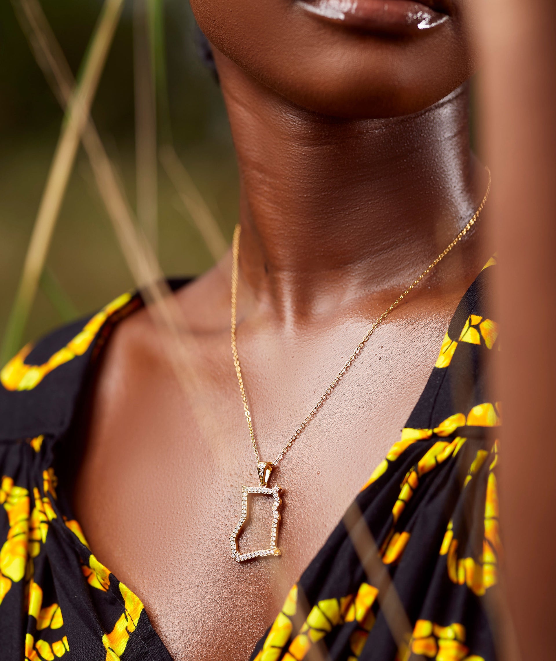 A close-up of a model wearing CASA London's 18k gold plated Ghana Pavé Necklace. The delicate gold chain features a pendant shaped like the country of Ghana, outlined with shimmering pavé-set stones. The model wears a black garment with a vibrant yellow pattern, providing a bold and culturally rich backdrop that highlights the brand's commitment to sophisticated luxury.