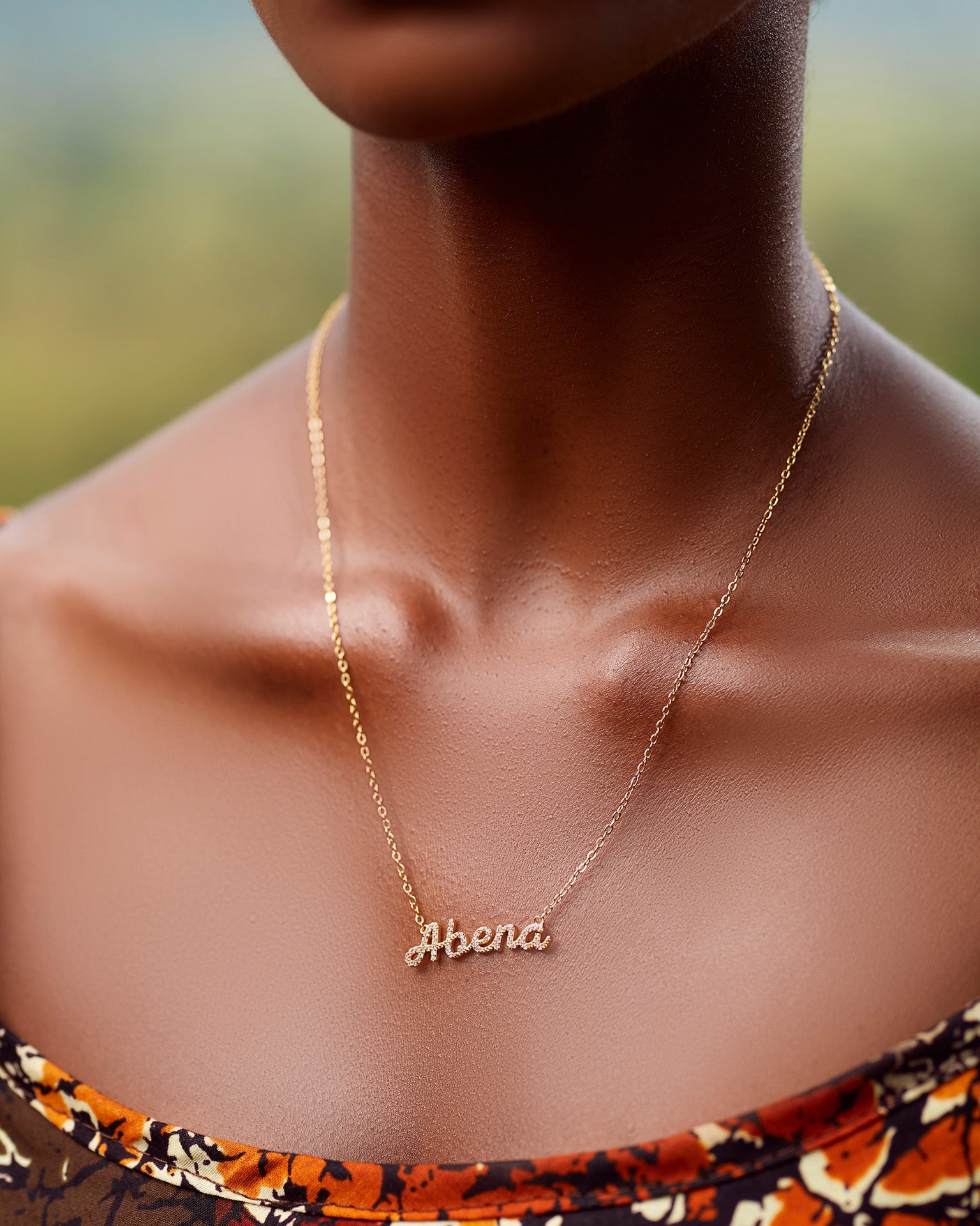 A close-up of a model wearing CASA London's 18k gold Abena Pavé Necklace. The delicate gold chain features a pendant with the name "Abena" intricately encrusted with shimmering pavé-set stones, celebrating the tradition of those born on a Tuesday. The model wears a patterned garment, providing a sophisticated backdrop that highlights the brand's commitment to elegant, narrative-driven luxury.