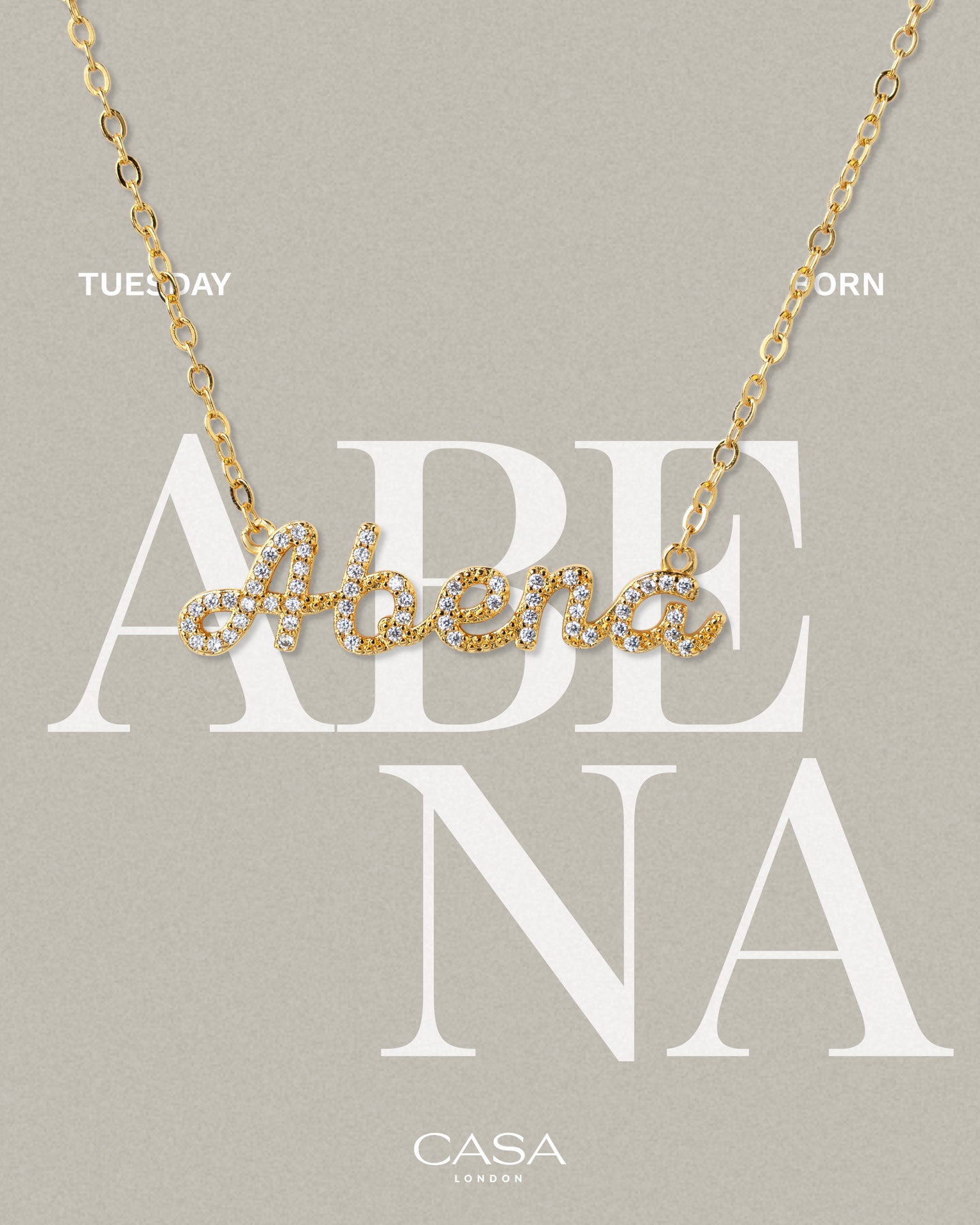 A product shot of CASA London’s 18k gold Abena Pavé Necklace. The delicate gold chain features a script pendant spelling "Abena," encrusted with shimmering pavé-set stones against a neutral, textured background. Overlaid text reads "TUESDAY BORN," highlighting the brand's commitment to celebrating cultural heritage and personal narratives through modern luxury.