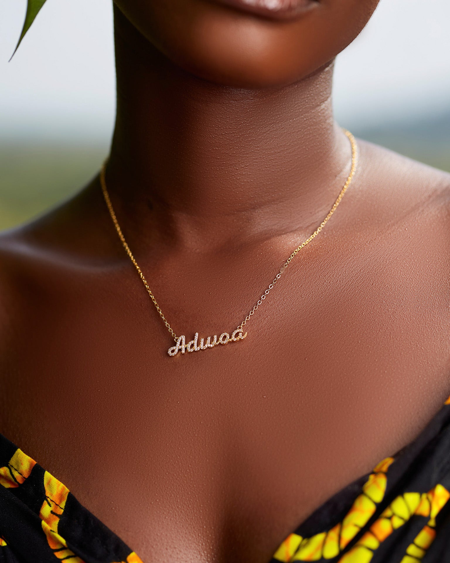 A close-up of a model wearing CASA London's 18k gold Adwoa Pavé Necklace. The delicate gold chain features a pendant with the name "Adwoa" intricately encrusted with shimmering pavé-set stones, celebrating the tradition of those born on a Monday. The model wears a black garment with a vibrant yellow pattern, providing a bold backdrop that highlights the brand's commitment to elegant, narrative-driven luxury.