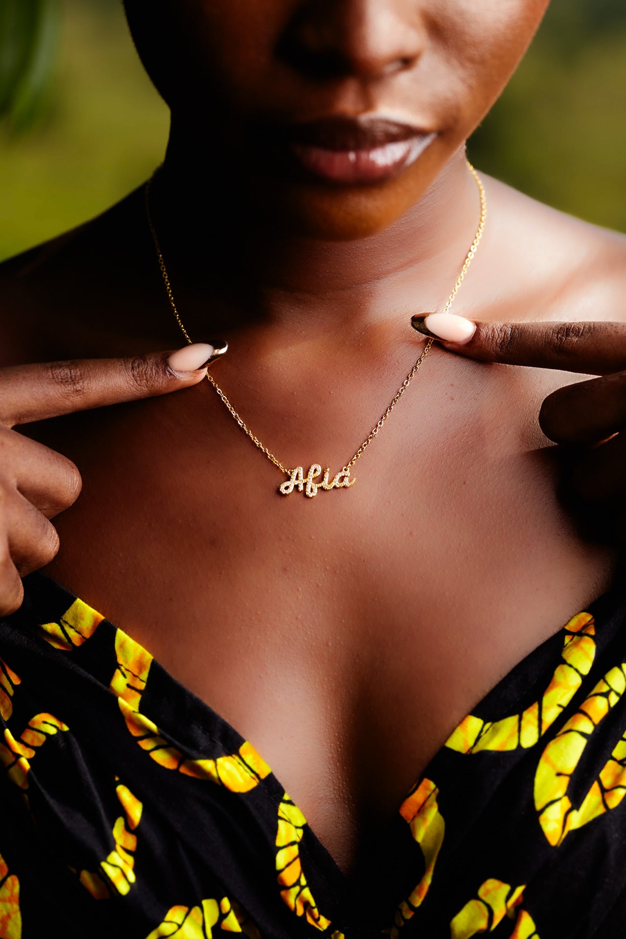 A close-up of a model wearing CASA London's 18k gold plated Afia Pavé Necklace. The delicate gold chain features a script pendant spelling "Afia" intricately encrusted with shimmering pavé-set stones, celebrating the tradition of those born on a Friday. The model wears a black garment with a vibrant yellow pattern, providing a bold backdrop that highlights the brand's commitment to elegant, narrative-driven luxury.