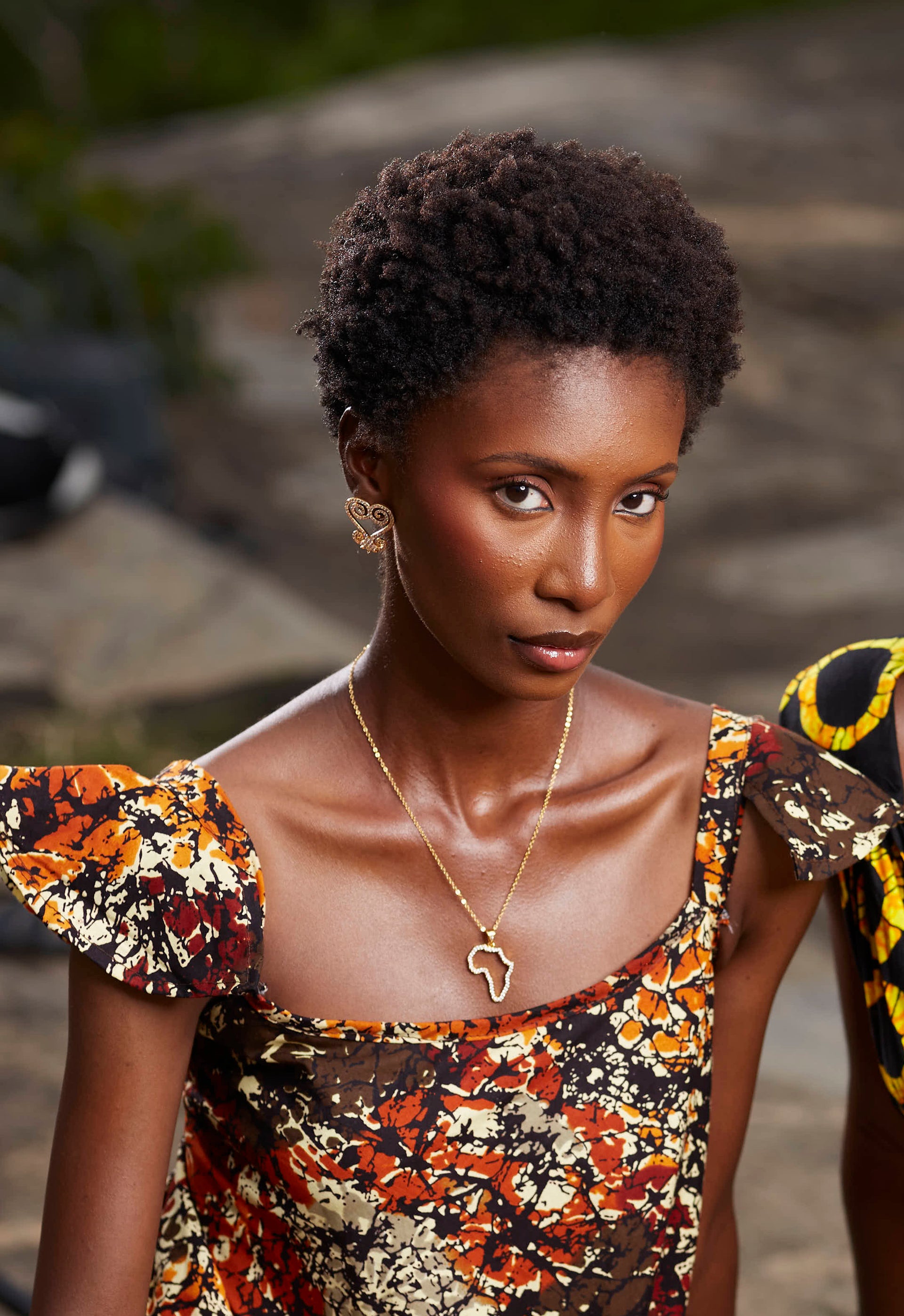 A portrait of a model wearing CASA London’s 18k gold plated Africa Pavé Necklace paired with gold Sankofa Pavé earrings. The necklace features a delicate gold chain with a pendant shaped like the continent of Africa, meticulously outlined with shimmering pavé-set stones. The model wears a patterned, earthy-toned garment with ruffled sleeves, and the natural outdoor lighting highlights the brand's commitment to blending sophisticated luxury with cultural storytelling.