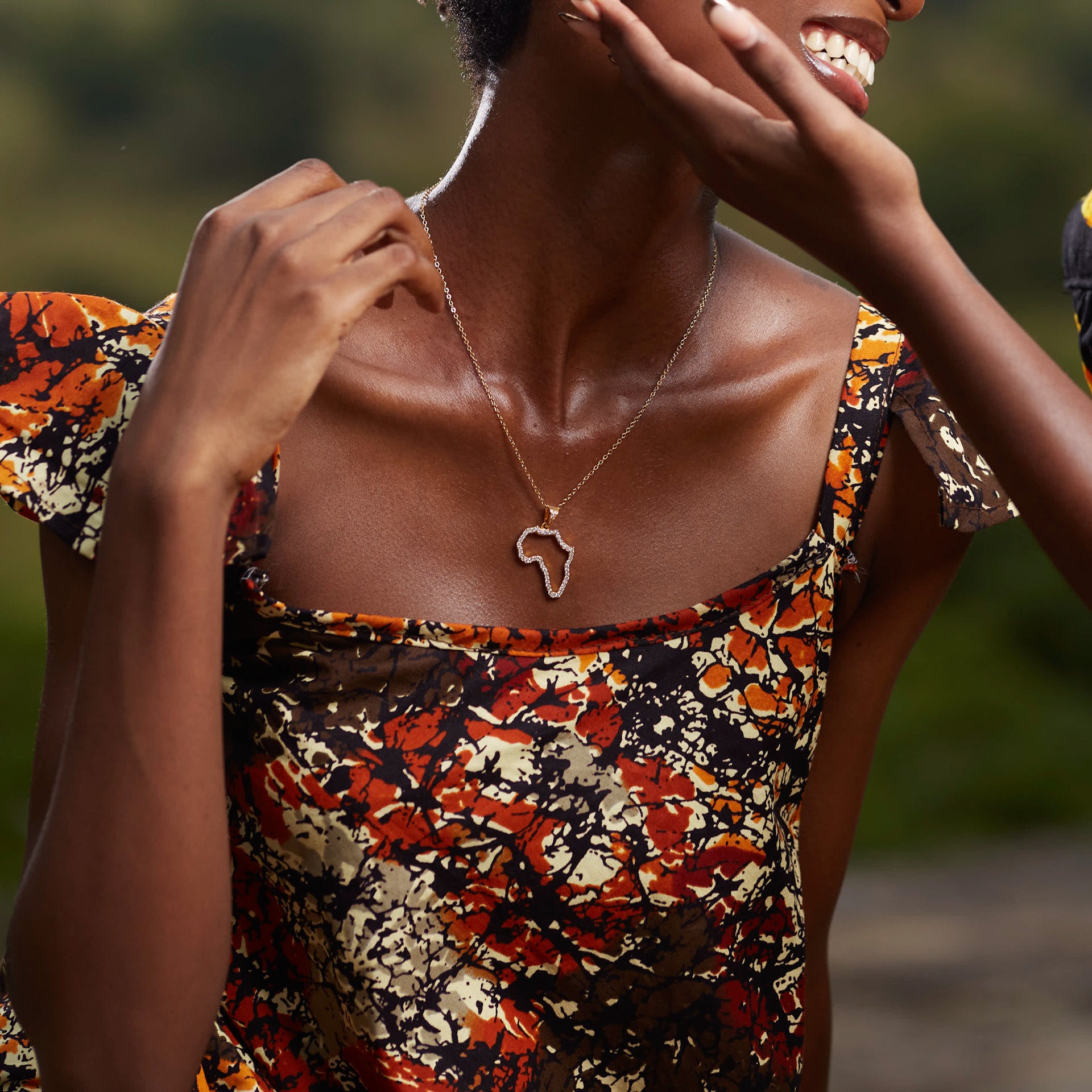 A portrait of a model wearing CASA London’s 18k gold plated Africa Pavé Necklace paired with gold Sankofa Pavé earrings. The necklace features a delicate gold chain with a pendant shaped like the continent of Africa, meticulously outlined with shimmering pavé-set stones. The model wears a patterned, earthy-toned garment with ruffled sleeves, and the natural outdoor lighting highlights the brand's commitment to blending sophisticated luxury with cultural storytelling.