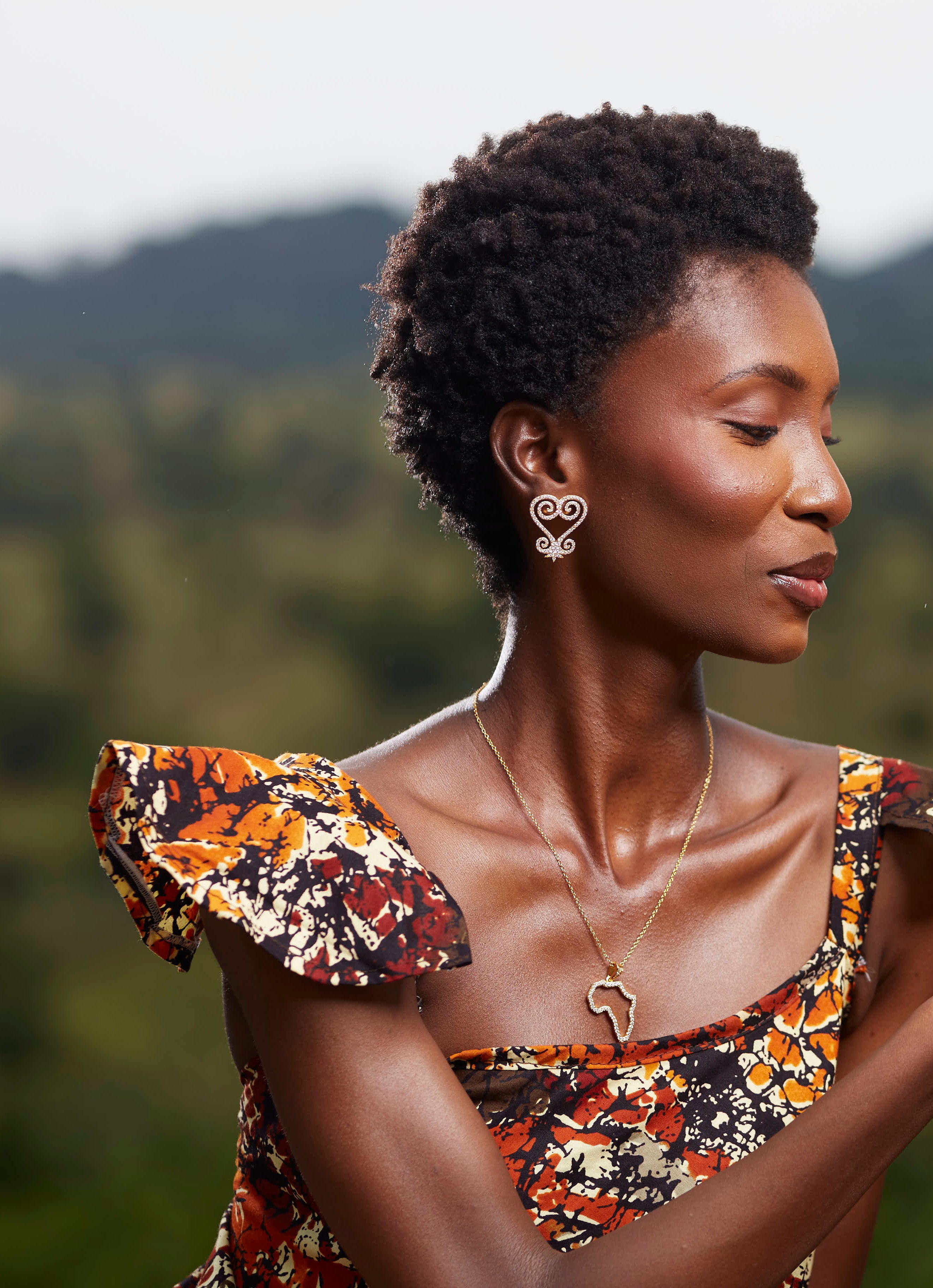 A portrait of a model wearing CASA London’s 18k gold plated Africa Pavé Necklace paired with gold Sankofa Pavé earrings. The necklace features a delicate gold chain with a pendant shaped like the continent of Africa, meticulously outlined with shimmering pavé-set stones. The model wears a patterned, earthy-toned garment with ruffled sleeves, and the natural outdoor lighting highlights the brand's commitment to blending sophisticated luxury with cultural storytelling.