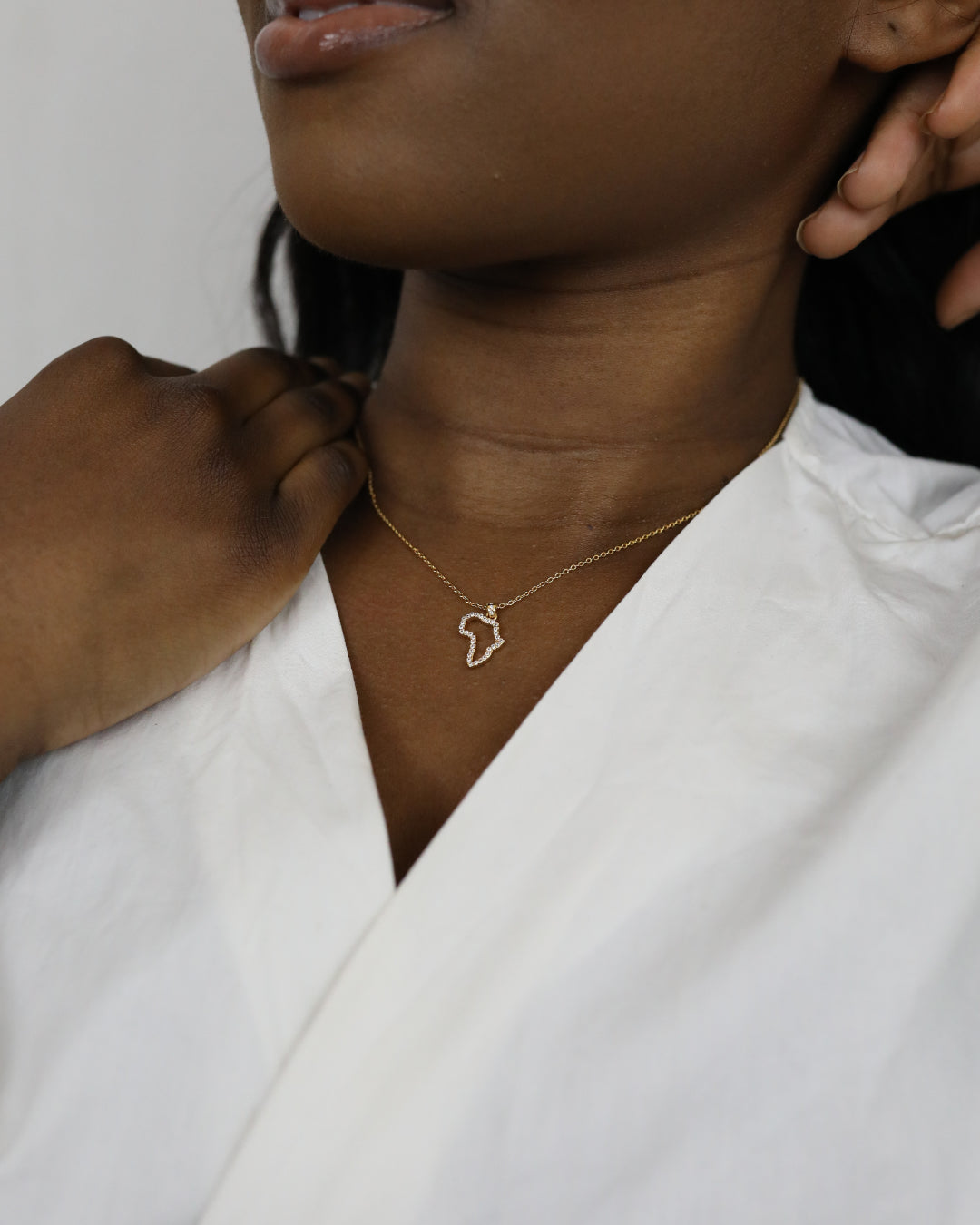 A close-up of a model wearing CASA London's 18k gold plated Mini Africa Pavé Necklace. The delicate gold chain features a pendant shaped like the continent of Africa, outlined with shimmering pavé-set stones. The model wears a white, V-neck garment, providing a clean and elegant backdrop for the necklace.
