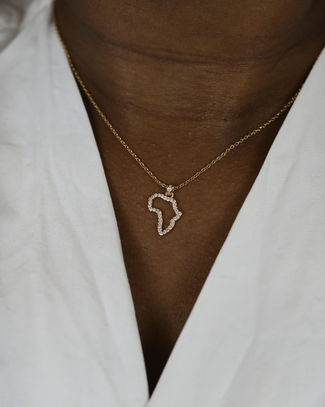 A close-up of a model wearing CASA London's 18k gold plated Mini Africa Pavé Necklace. The delicate gold chain features a pendant shaped like the continent of Africa, outlined with shimmering pavé-set stones. The model wears a white, V-neck garment, providing a clean and elegant backdrop that highlights the brand's commitment to sophisticated luxury.