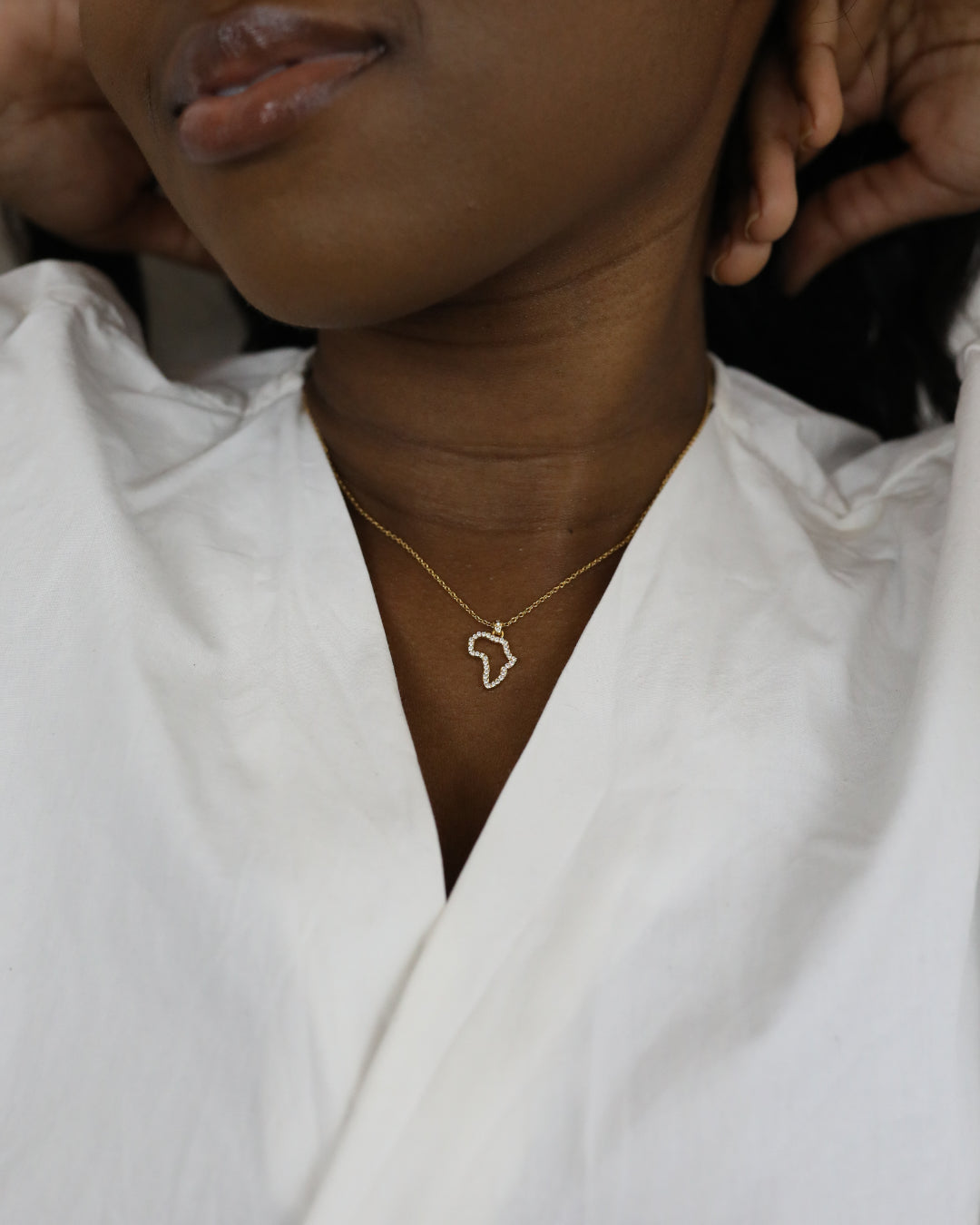 A close-up of a model wearing CASA London's 18k gold plated Mini Africa Pavé Necklace. The delicate gold chain features a pendant shaped like the continent of Africa, outlined with shimmering pavé-set stones. The model wears a white, V-neck garment, providing a clean and elegant backdrop that highlights the brand's commitment to sophisticated luxury.