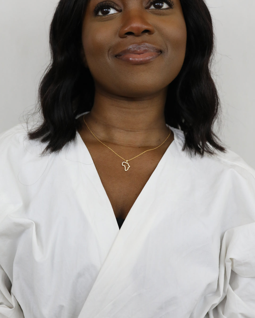 A portrait of a model wearing CASA London's 18k gold plated Mini Africa Pavé Necklace. The delicate gold chain features a pendant shaped like the continent of Africa, outlined with shimmering pavé-set stones. The model wears a white, V-neck garment, providing a clean and elegant backdrop for the necklace.