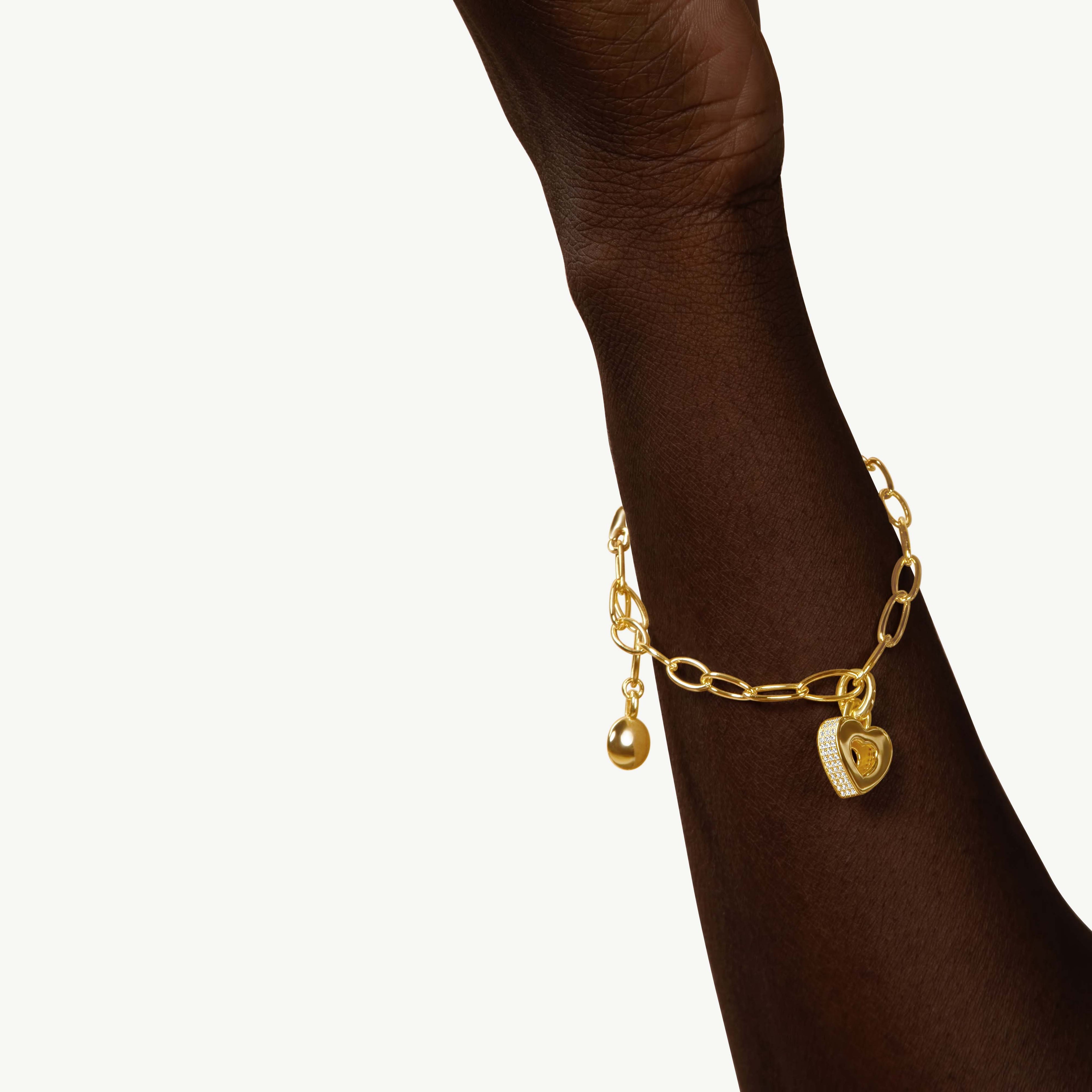 CASA LONDON Akoma Pavé Charm in 18ct gold vermeil worn on a dark-toned wrist — heart-shaped Adinkra-inspired charm with white zirconia pavé stones on a gold link bracelet