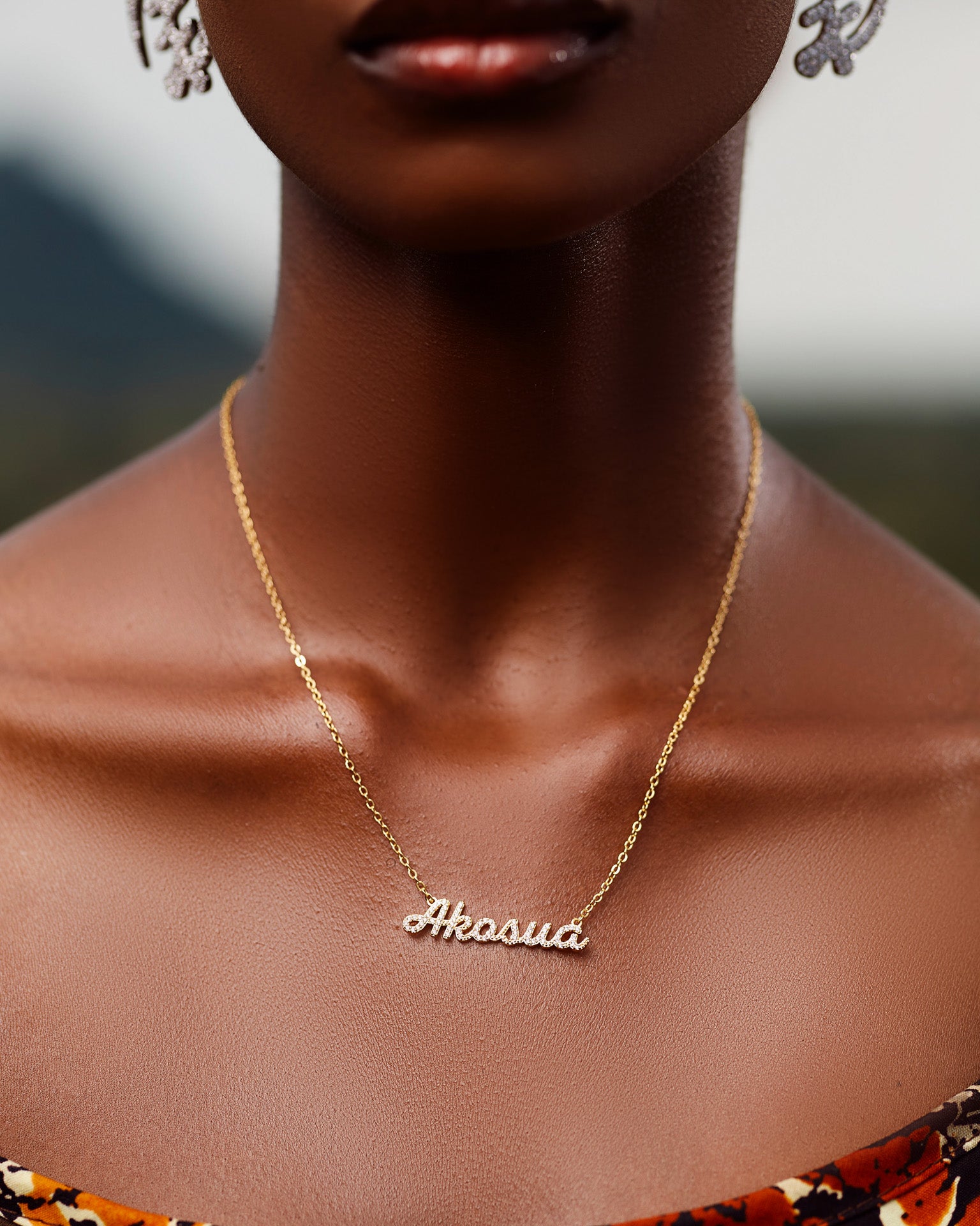 A close-up of a model wearing CASA London's 18k gold Akosua Pavé Necklace. The delicate gold chain features a pendant with the name "Akosua" intricately encrusted with shimmering pavé-set stones, celebrating the tradition of those born on a Sunday. The model wears a patterned garment, providing a sophisticated backdrop that highlights the brand's commitment to elegant, narrative-driven luxury.