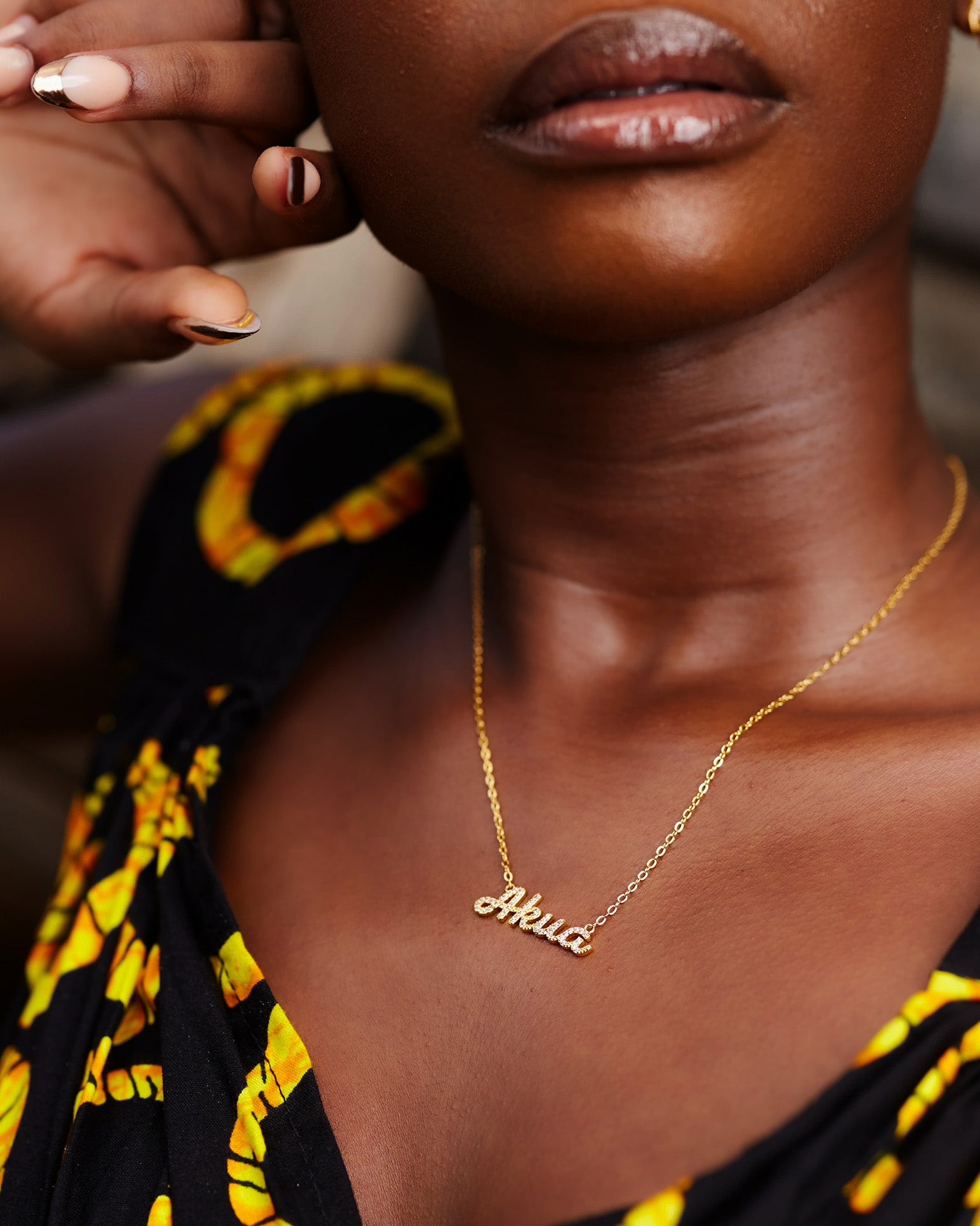A close-up of a model wearing CASA London's 18k gold plated Akua Pavé Necklace. The delicate gold chain features a pendant with the name "Akua" intricately encrusted with shimmering pavé-set stones, celebrating the tradition of those born on a Wednesday. The model wears a black garment with a vibrant yellow pattern, providing a bold backdrop that highlights the brand's commitment to elegant, narrative-driven luxury.