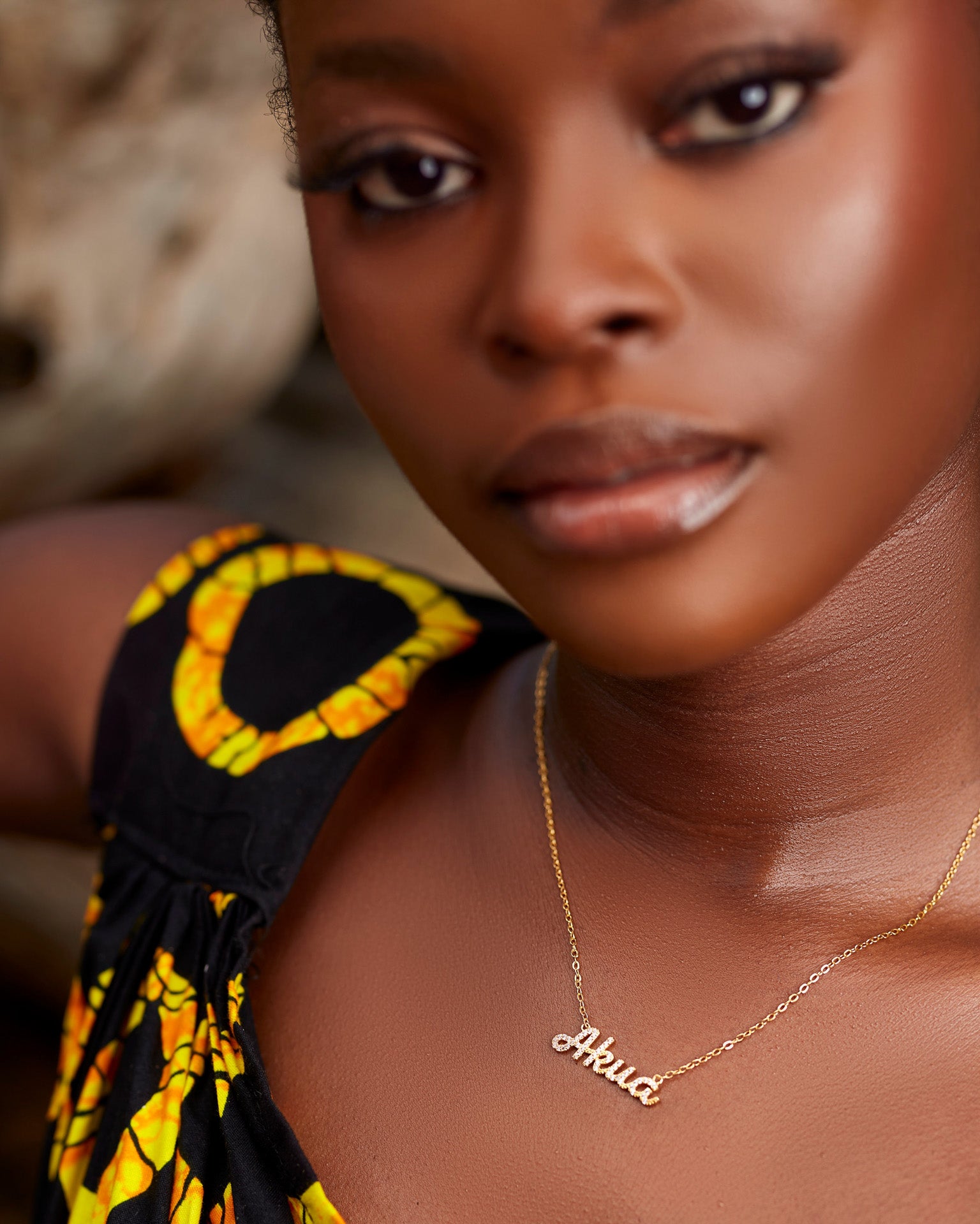A close-up of a model wearing CASA London's 18k gold plated Akua Pavé Necklace. The delicate gold chain features a pendant with the name "Akua" intricately encrusted with shimmering pavé-set stones, celebrating the tradition of those born on a Wednesday. The model wears a black garment with a vibrant yellow pattern, providing a bold backdrop that highlights the brand's commitment to elegant, narrative-driven luxury.