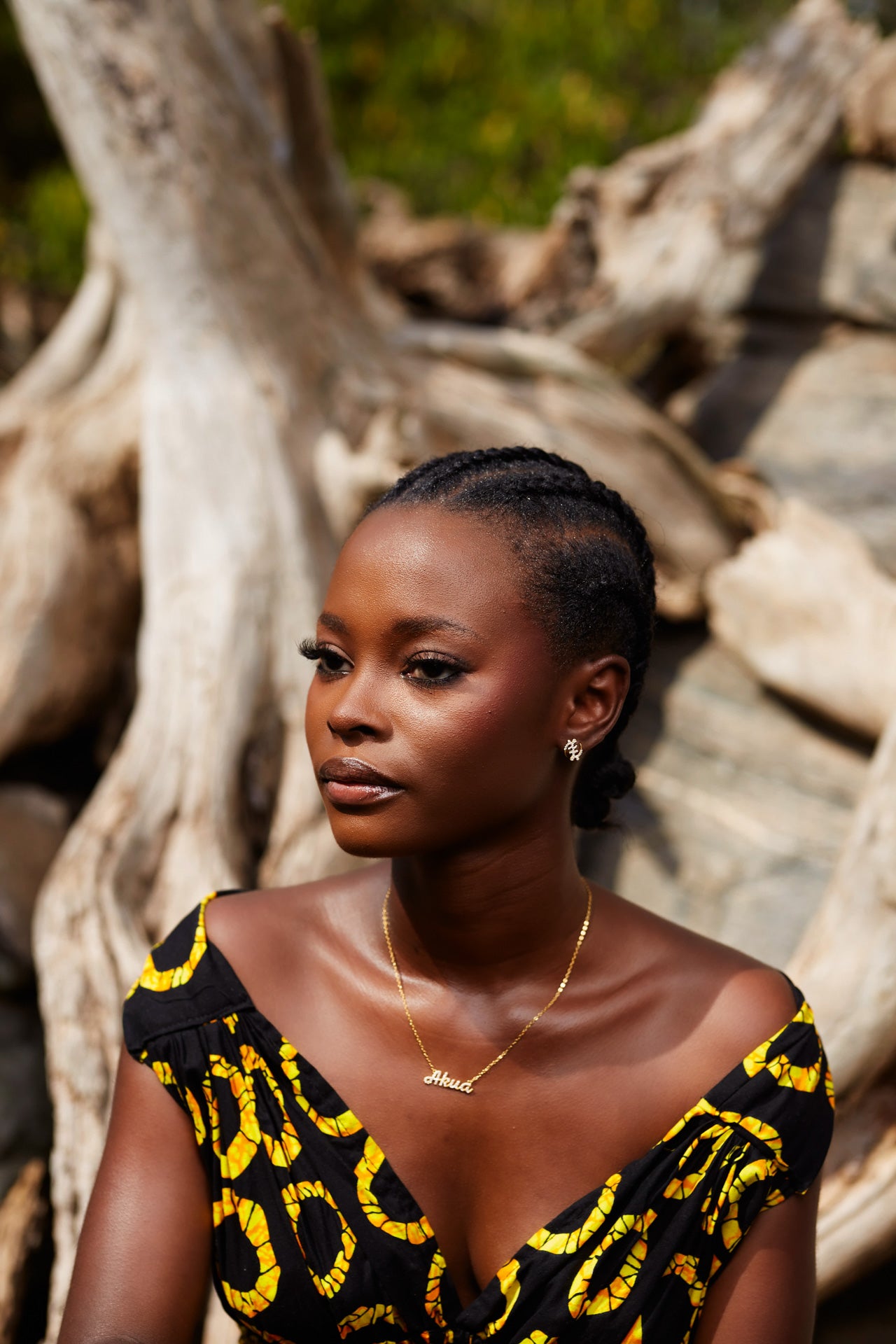 A portrait of a model wearing CASA London’s 18k gold plated Akua Pavé Necklace paired with coordinating pavé stud earrings. The necklace features a delicate gold chain with a script pendant spelling "Akua," intricately encrusted with shimmering pavé-set stones to celebrate those born on a Wednesday. The model’s hair is styled in intricate braids gathered into a low bun, and she wears a black garment with a vibrant yellow pattern. The rustic driftwood backdrop and natural lighting highlight the brand's commi