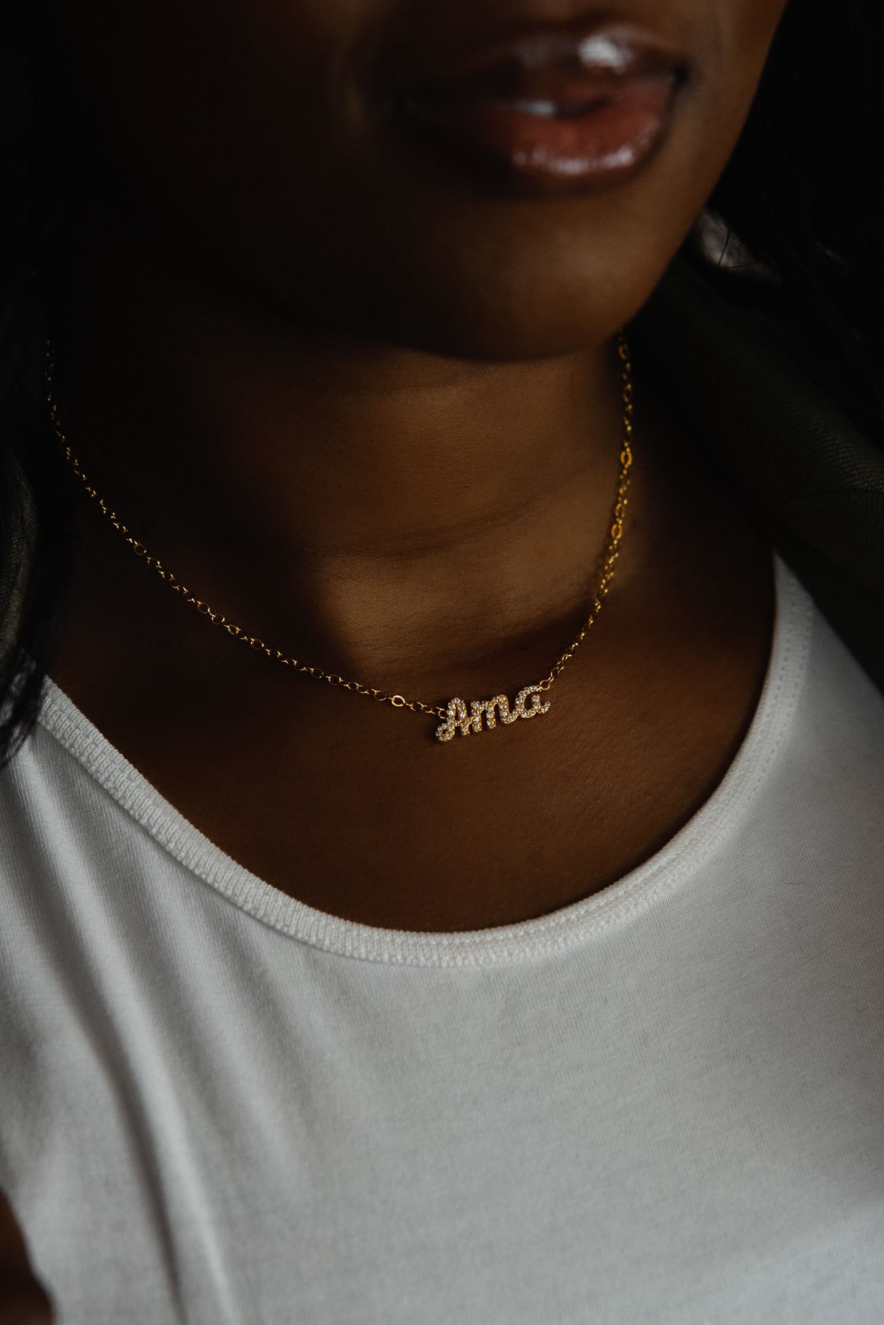 A close-up of a model wearing CASA London's 18k gold Ama Pavé Necklace. The delicate gold chain features a pendant with the name "Ama" intricately encrusted with shimmering pavé-set stones, celebrating the tradition of those born on a Saturday. The model wears a white top, providing a clean backdrop that highlights the brand's commitment to elegant, narrative-driven luxury.