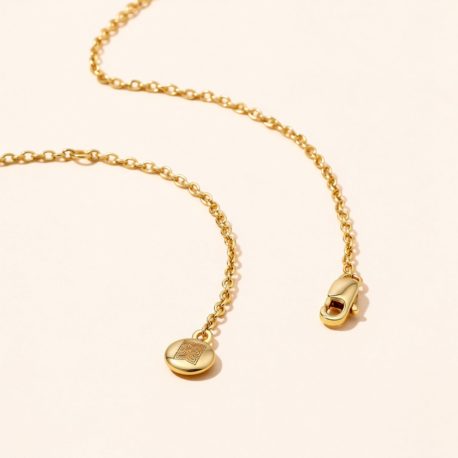 Flat lay close-up of the CASA London Custom Pavé Name Necklace chain in 18ct gold vermeil on S925 sterling silver, showing the lobster clasp and engraved CASA London brand insignia disc charm on a cream background