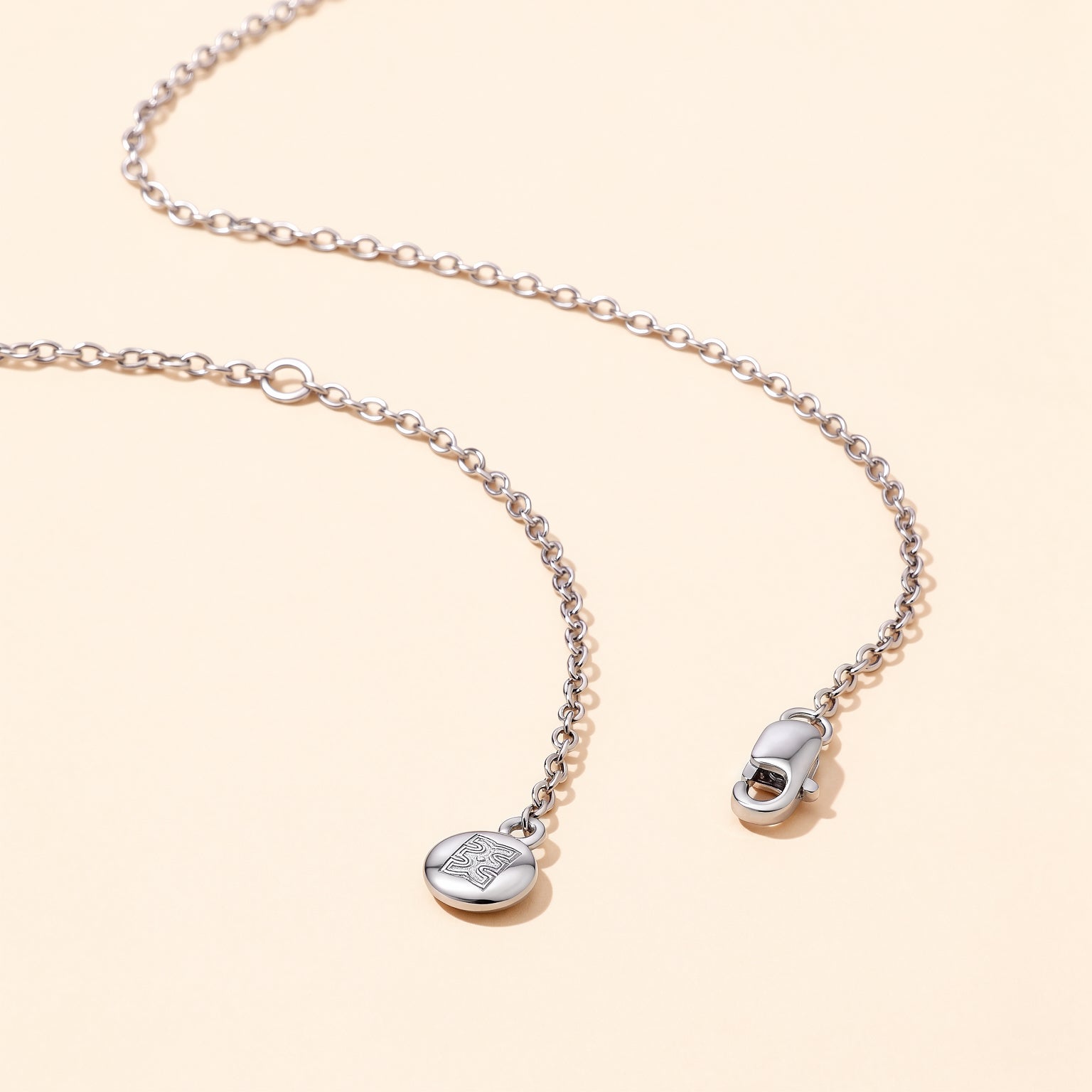 Close-up flat lay of the CASA London Custom Pavé Name Necklace chain in rhodium silver on S925 sterling silver, showing the lobster clasp and engraved CASA London brand insignia disc charm on a cream background