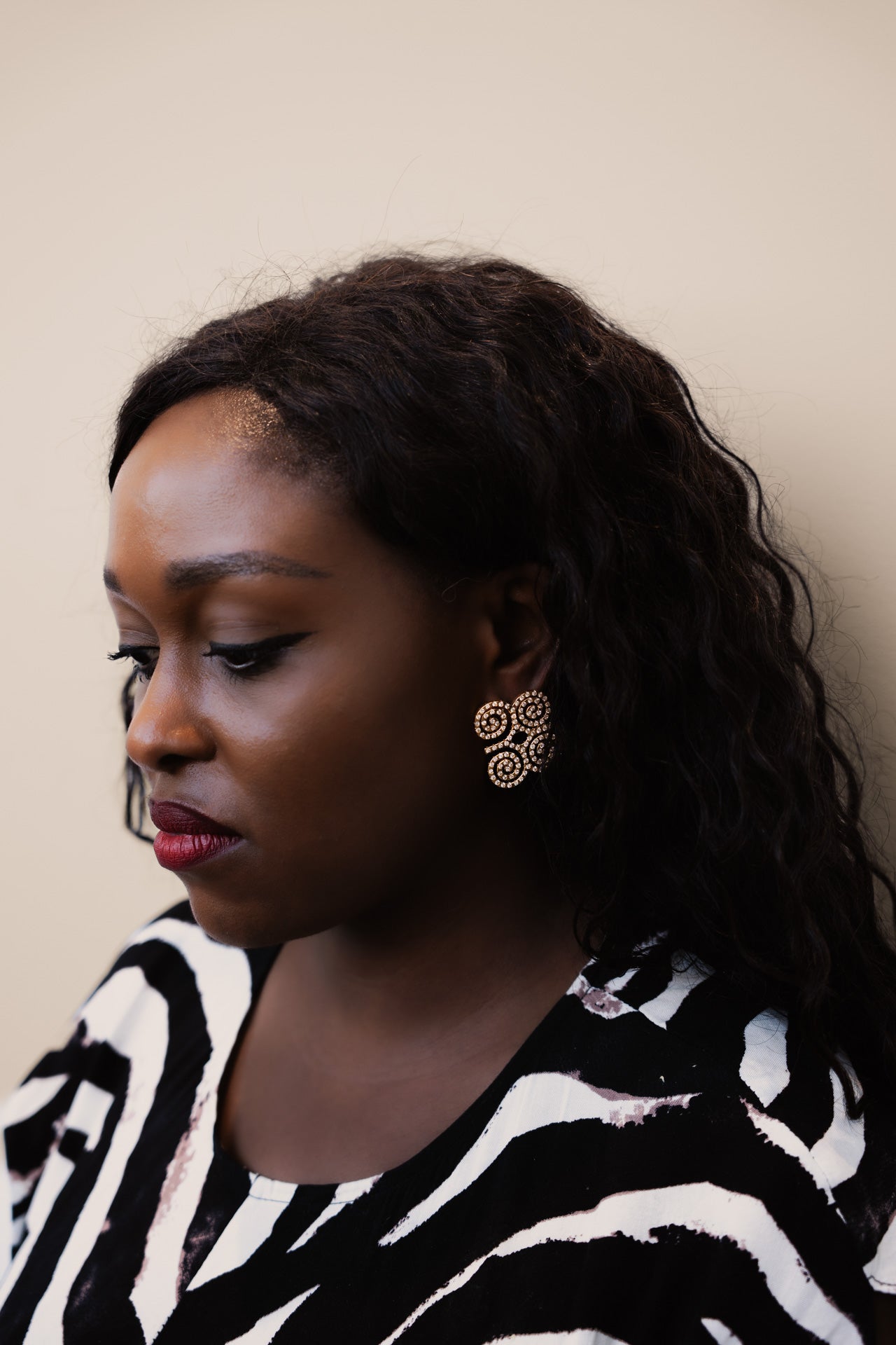 A close-up profile shot of a model wearing CASA London’s gold Dwennimmen Pavé Earrings. The intricate earrings are shaped into the Adinkra symbol of rams' horns—representing strength and humility—and are meticulously encrusted with shimmering pavé-set stones. The detailed shimmer against the soft-focus backdrop highlights the brand's commitment to elegant, narrative-driven luxury.