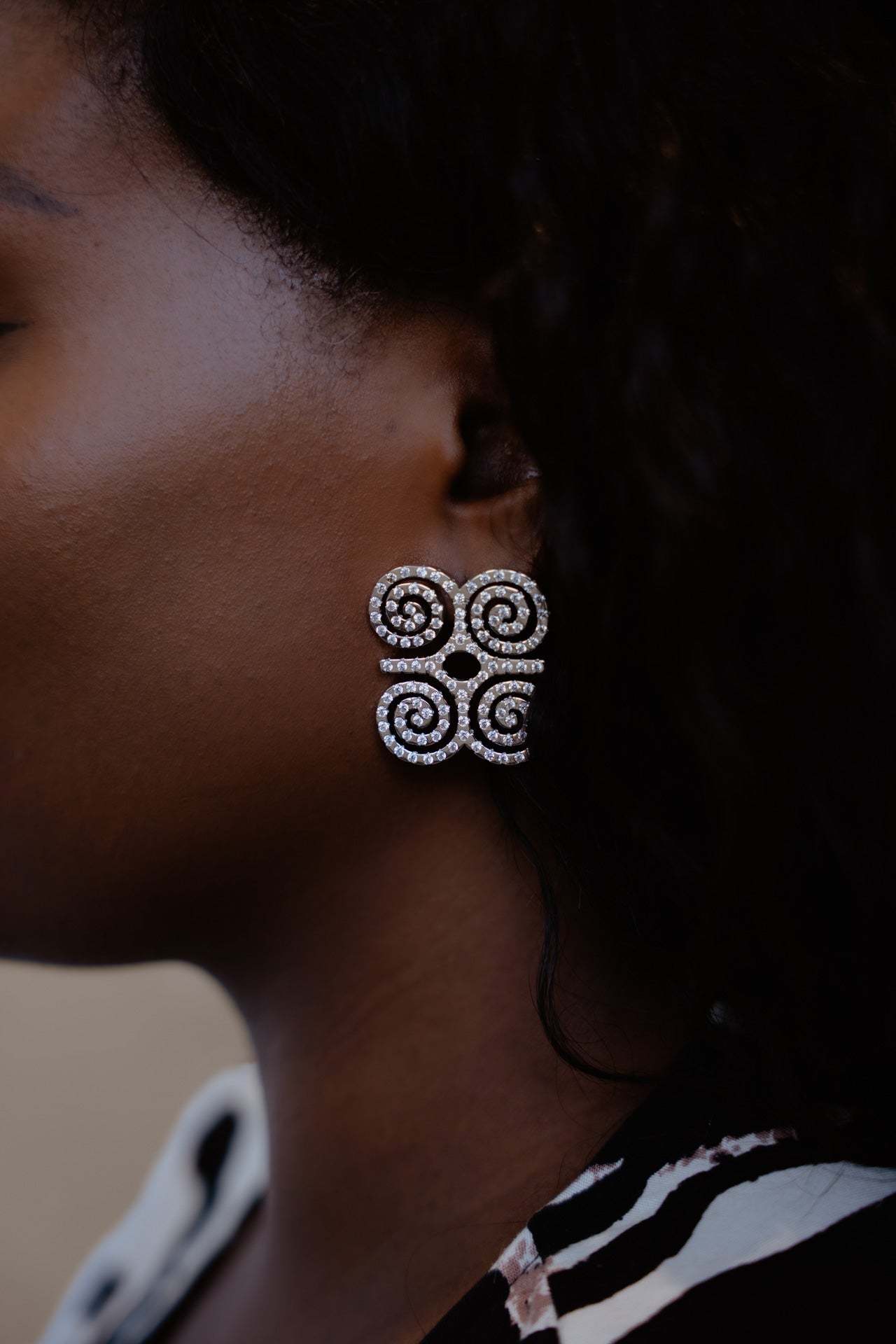 A close-up profile shot of a model wearing CASA London’s silver Dwennimmen Pavé Earrings. The intricate earrings are shaped into the Adinkra symbol of rams' horns—representing strength and humility—and are meticulously encrusted with shimmering pavé-set stones. The detailed shimmer against the soft-focus backdrop highlights the brand's commitment to elegant, narrative-driven luxury.