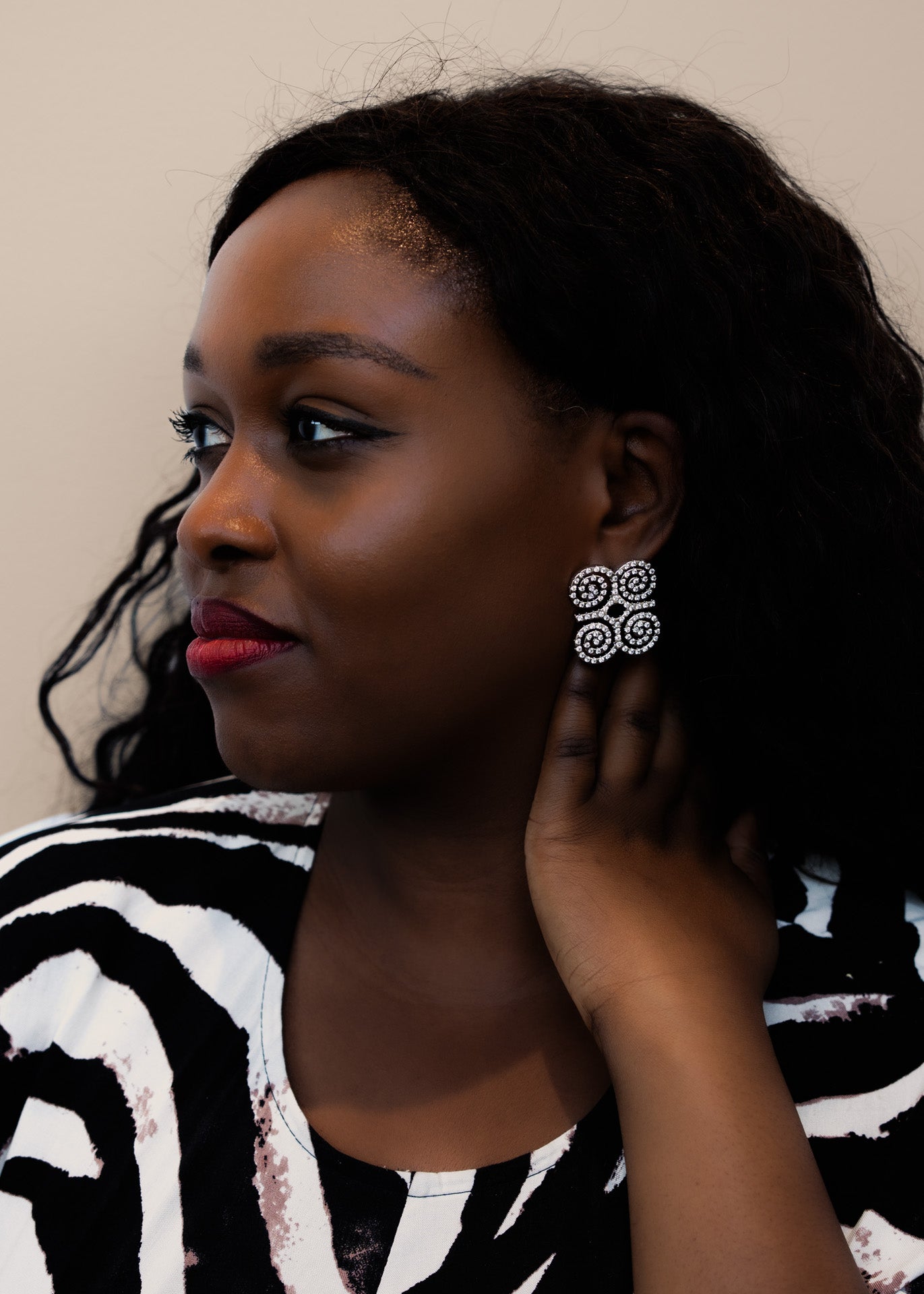 A close-up profile shot of a model wearing CASA London’s silver Dwennimmen Pavé Earrings. The intricate earrings are shaped into the Adinkra symbol of rams' horns—representing strength and humility—and are meticulously encrusted with shimmering pavé-set stones. The detailed shimmer against the soft-focus backdrop highlights the brand's commitment to elegant, narrative-driven luxury.