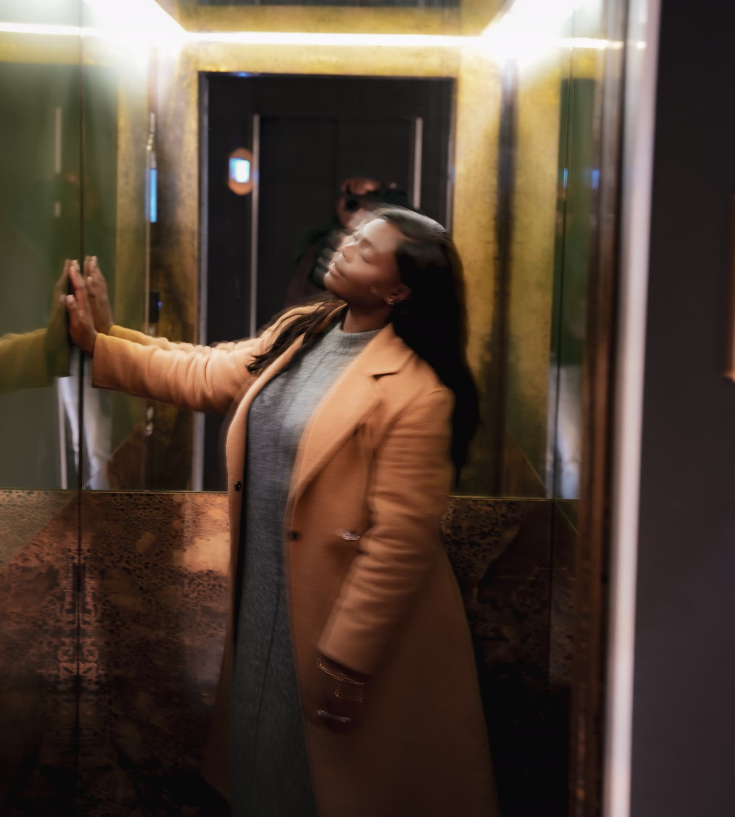Christine, founder of CASA London, resting one hand on the lift mirror with her head tilted back and her eyes closed, in a camel coat