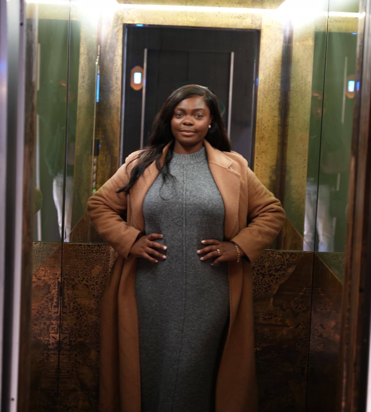 Christine, founder of CASA London, standing with her hands on her hips in a mirrored gold lift, wearing a camel coat, a grey dress and gold jewellery