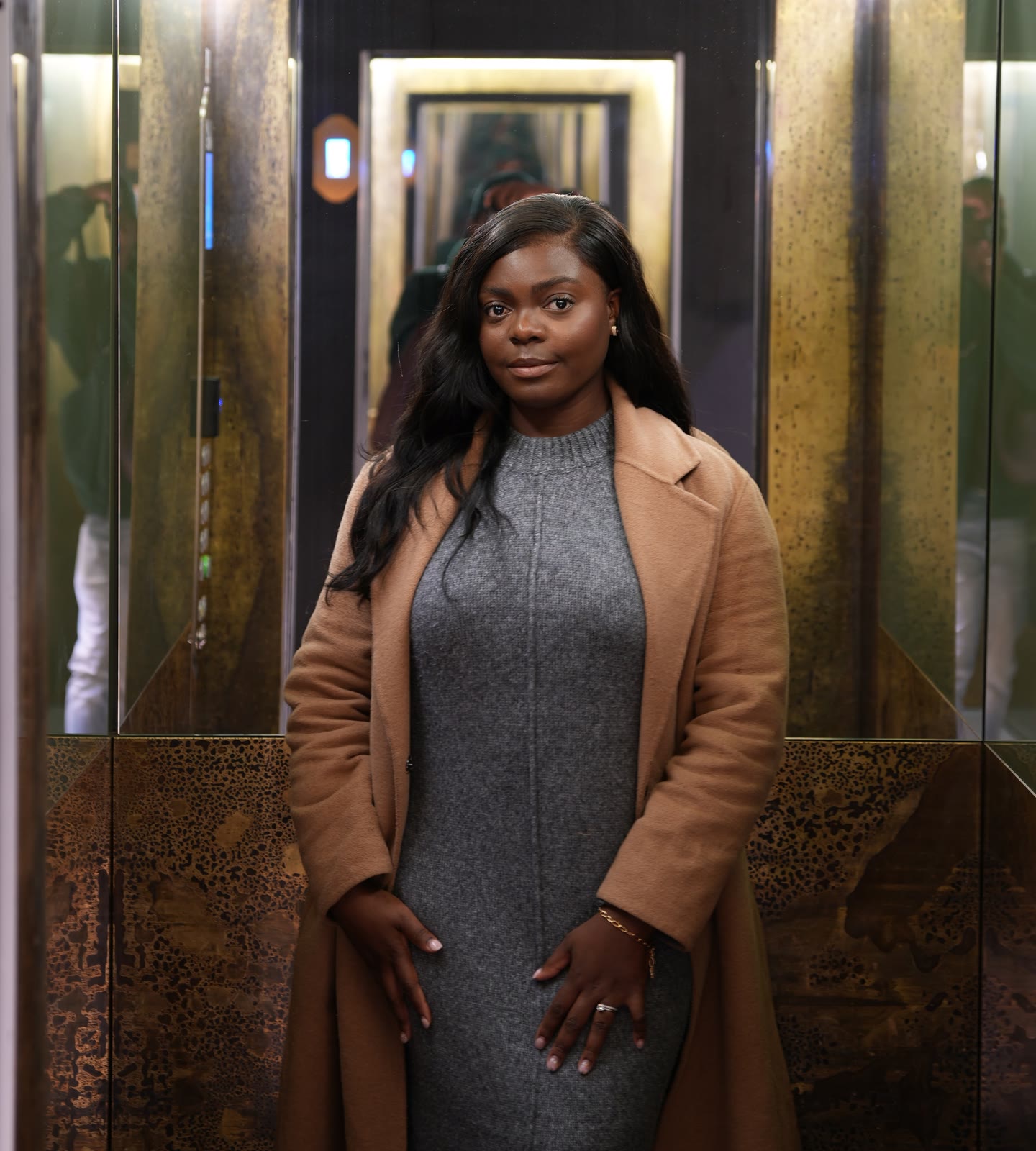Christine, founder of CASA London, standing in a gold-lit mirrored lift wearing a camel coat over a grey knit dress, looking calmly at the camera
