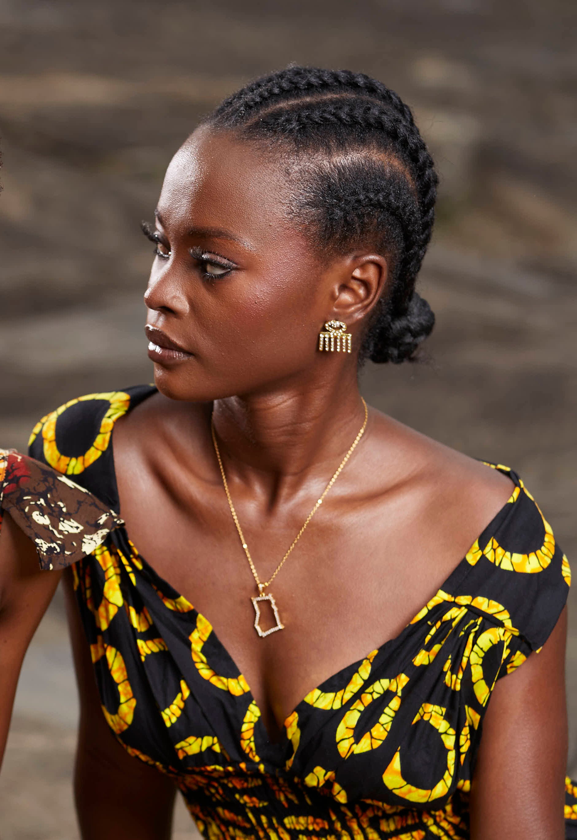 A portrait of a model wearing CASA London’s 18k gold plated Ghana Pavé Necklace paired with gold Duafe stud earrings. The necklace features a delicate gold chain with a pendant shaped like the country of Ghana, outlined with shimmering pavé-set stones. The model’s hair is styled in intricate braids gathered into a low bun, and she wears a black garment with a vibrant yellow pattern, providing a bold backdrop that highlights the brand's commitment to sophisticated luxury and cultural storytelling.