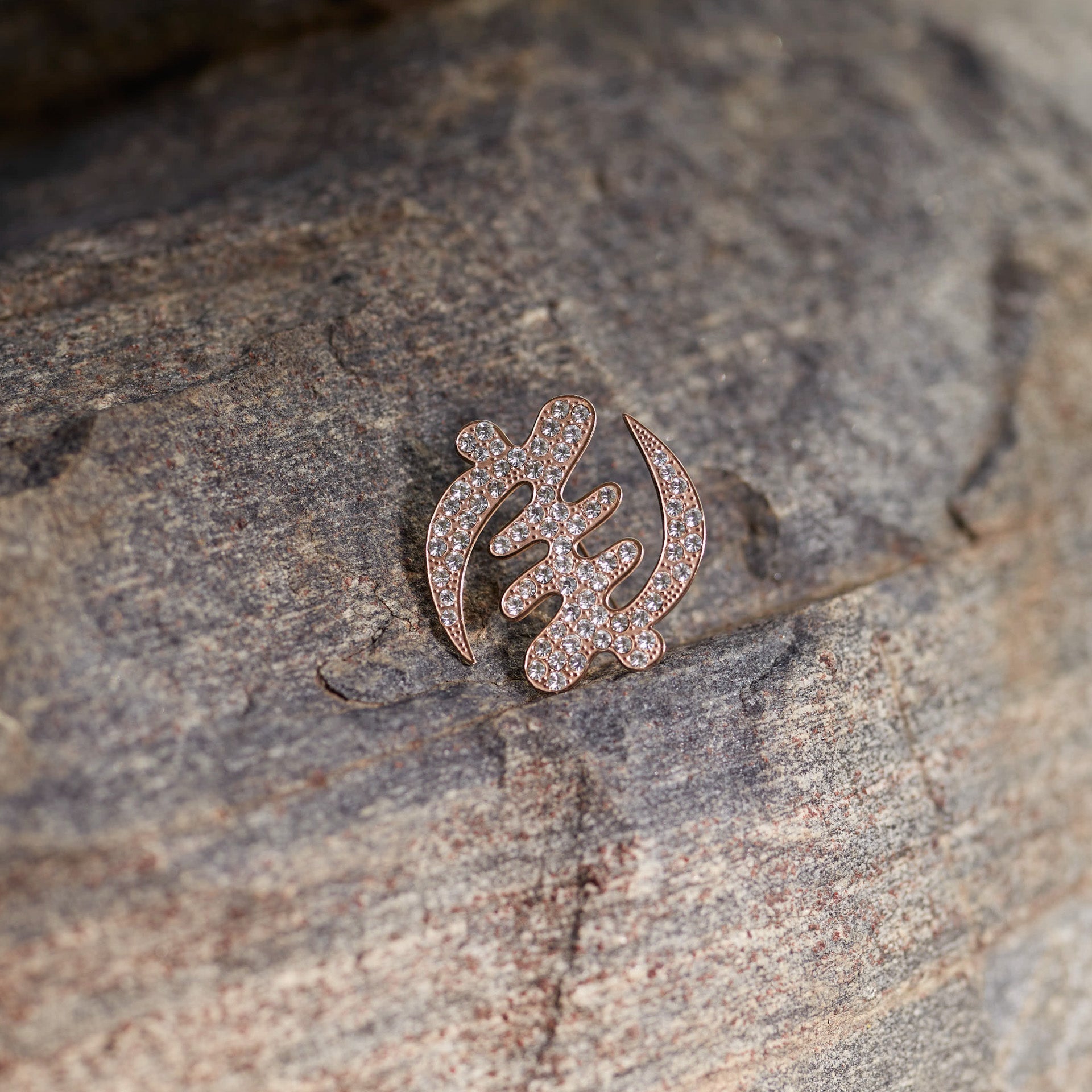 A detailed shot of CASA London's 18ct rose gold plated Gye Nyame lapel pin resting on the textured surface of a grey rock. The pin, meticulously shaped into the iconic Adinkra symbol and encrusted with shimmering pavé-set stones, catches the soft natural light. This outdoor setting highlights the brand's connection to heritage and its commitment to merging cultural significance with high-end luxury.