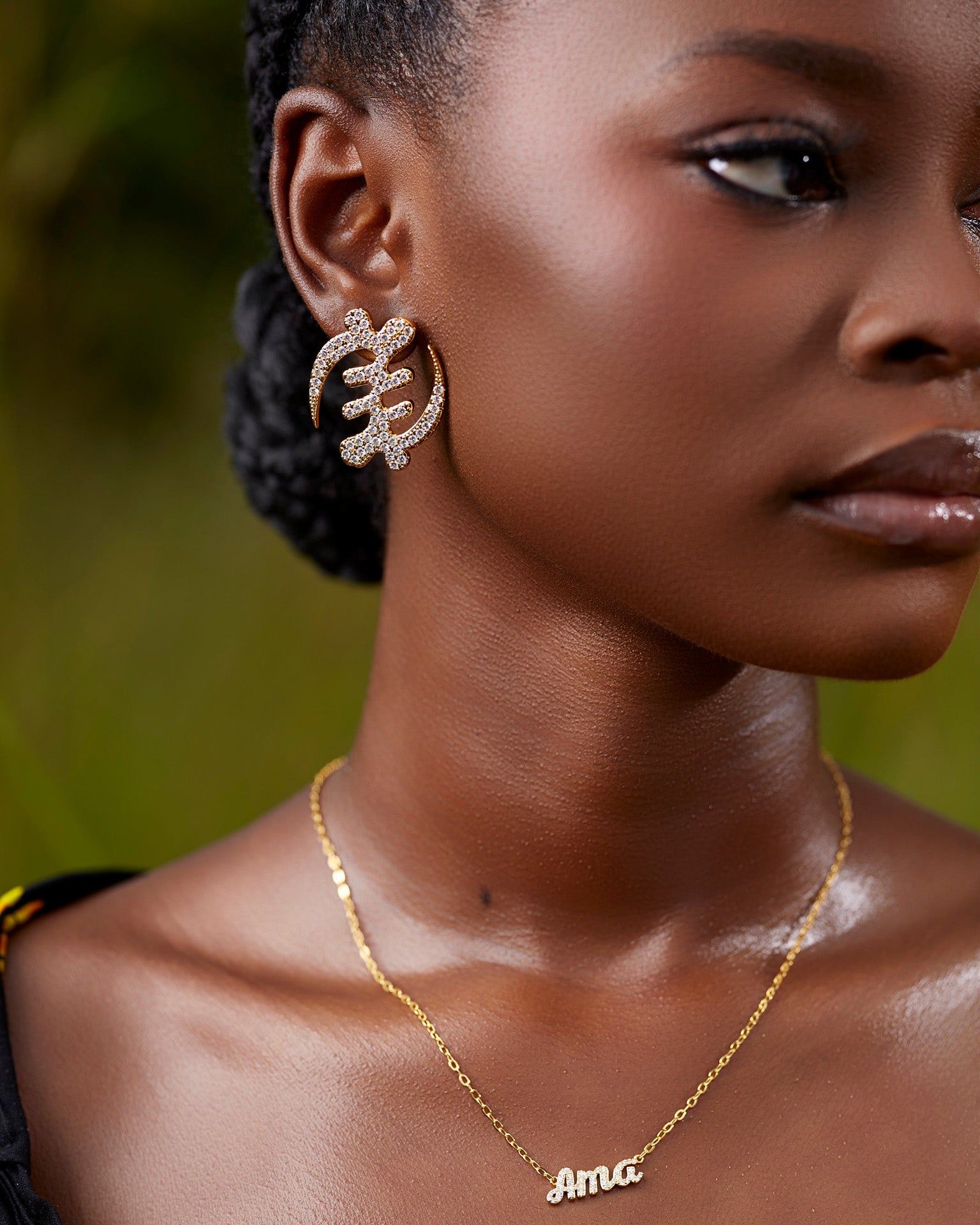Close-up of a model wearing a large gold Gye Nyame Adinkra symbol earring, featuring a bold pavé-set high-grade zircon design against a natural outdoor background.