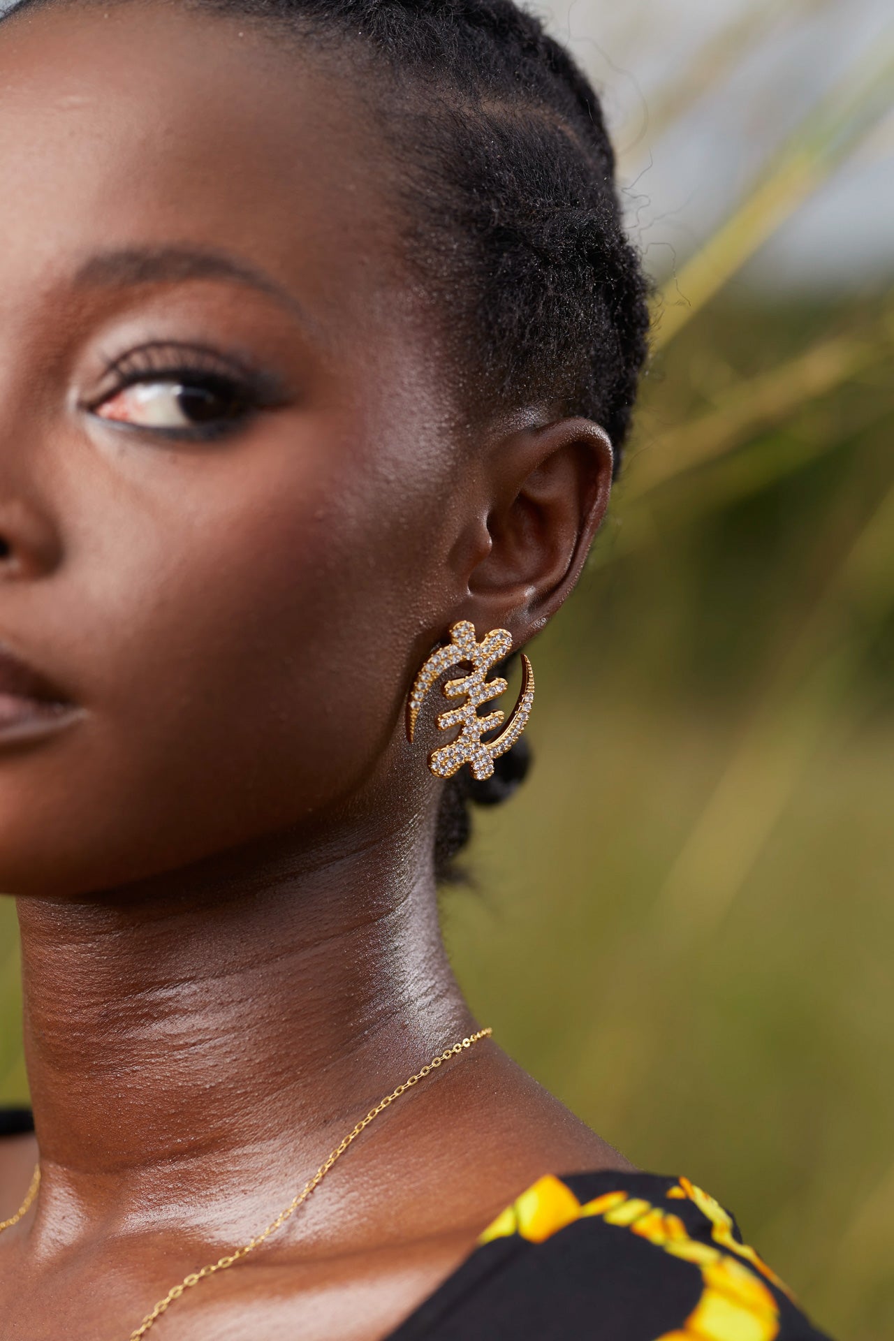 Close-up of a model wearing a large gold Gye Nyame Adinkra symbol earring, featuring a bold pavé-set high-grade zircon design against a natural outdoor background.