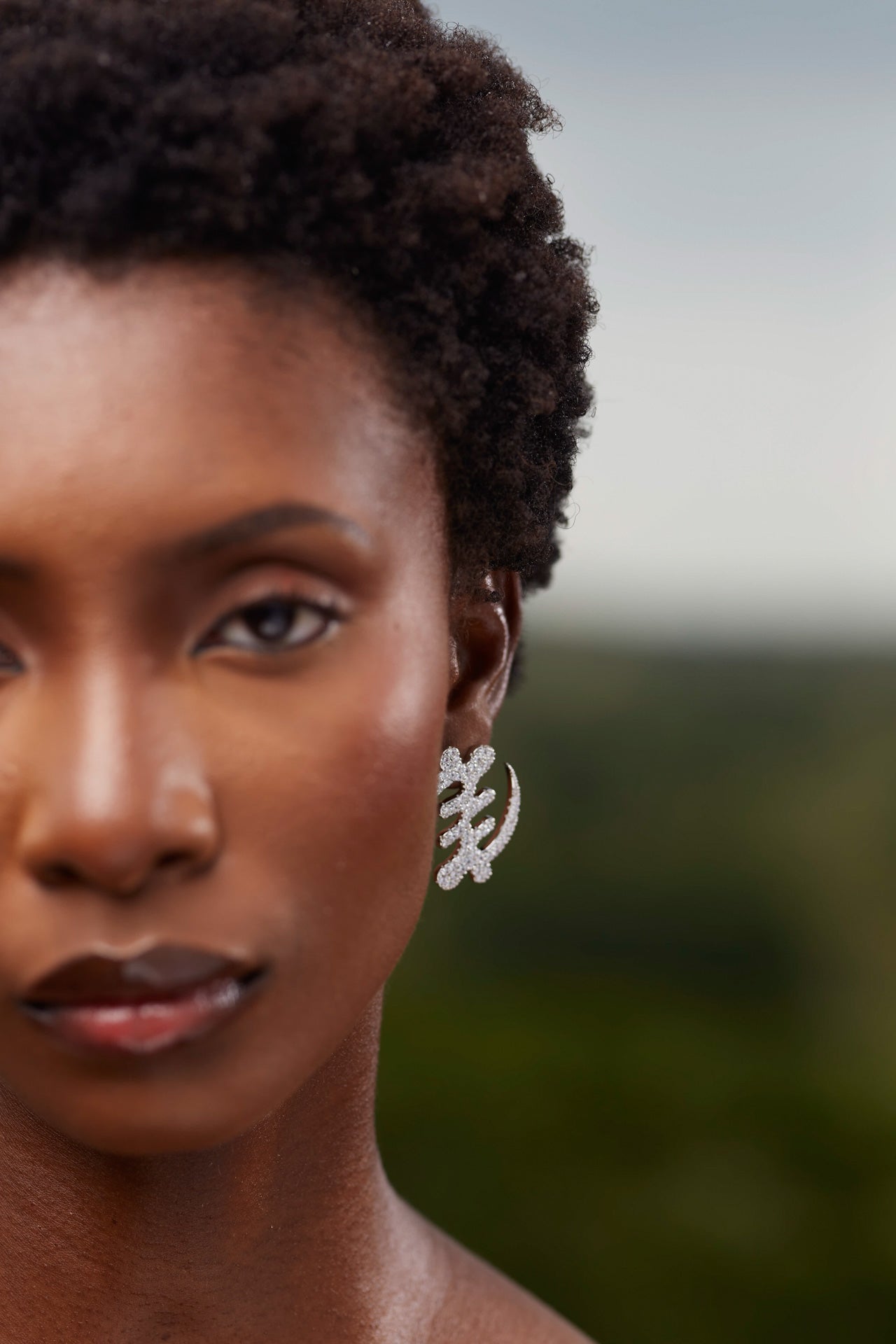 Model wearing large silver Gye Nyame Adinkra symbol earrings set with high-grade zircon stones, showcasing the traditional Ghanaian symbol of 'Except for God' in a shimmering pavé design.