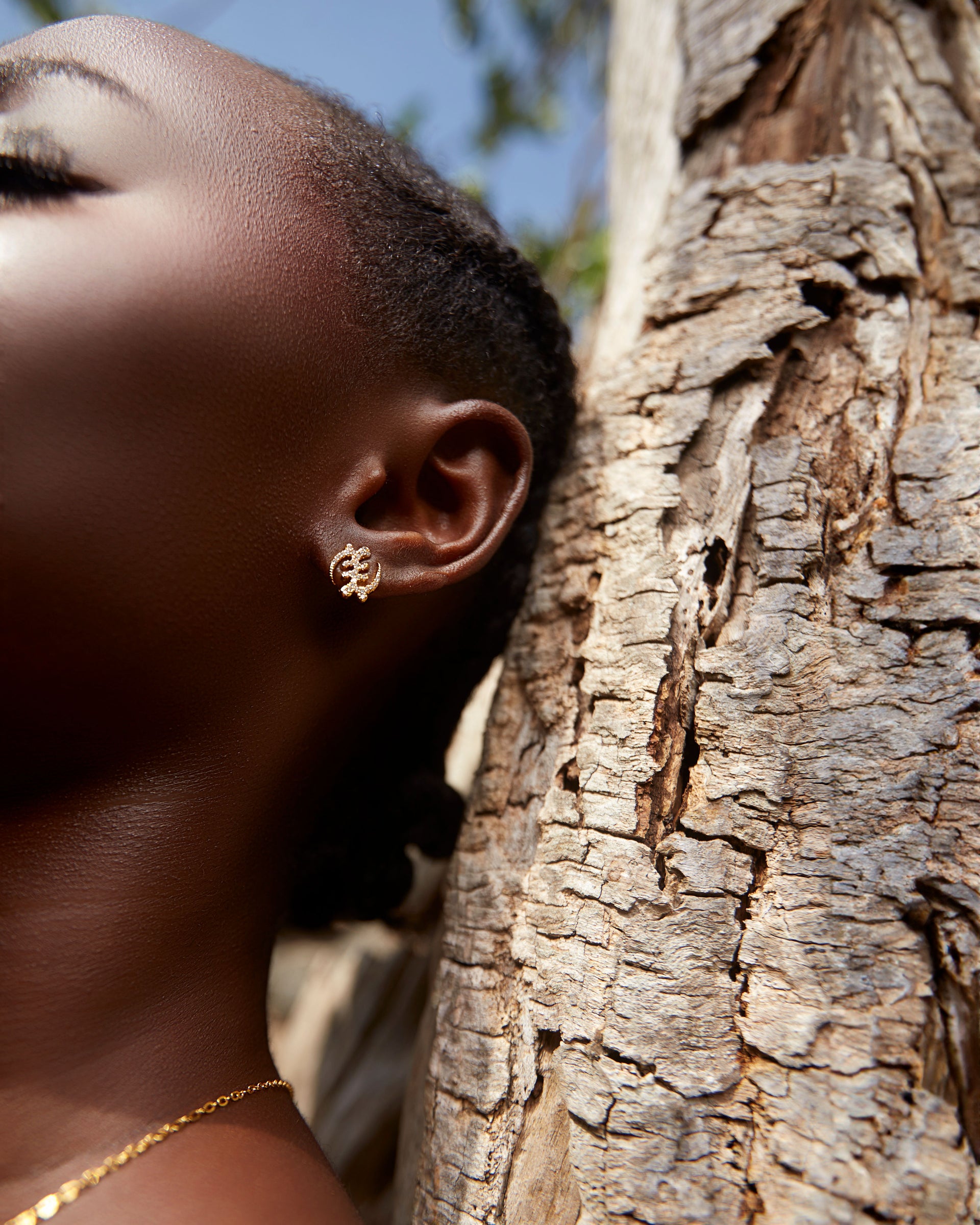 Close-up portrait of a model leaning against a textured tree trunk, wearing the CASA London Gye Nyame Pavé Earrings Mini. The gold stud is intricately shaped like the traditional Gye Nyame symbol and encrusted with shimmering pavé zircon stones, catching the natural outdoor light.