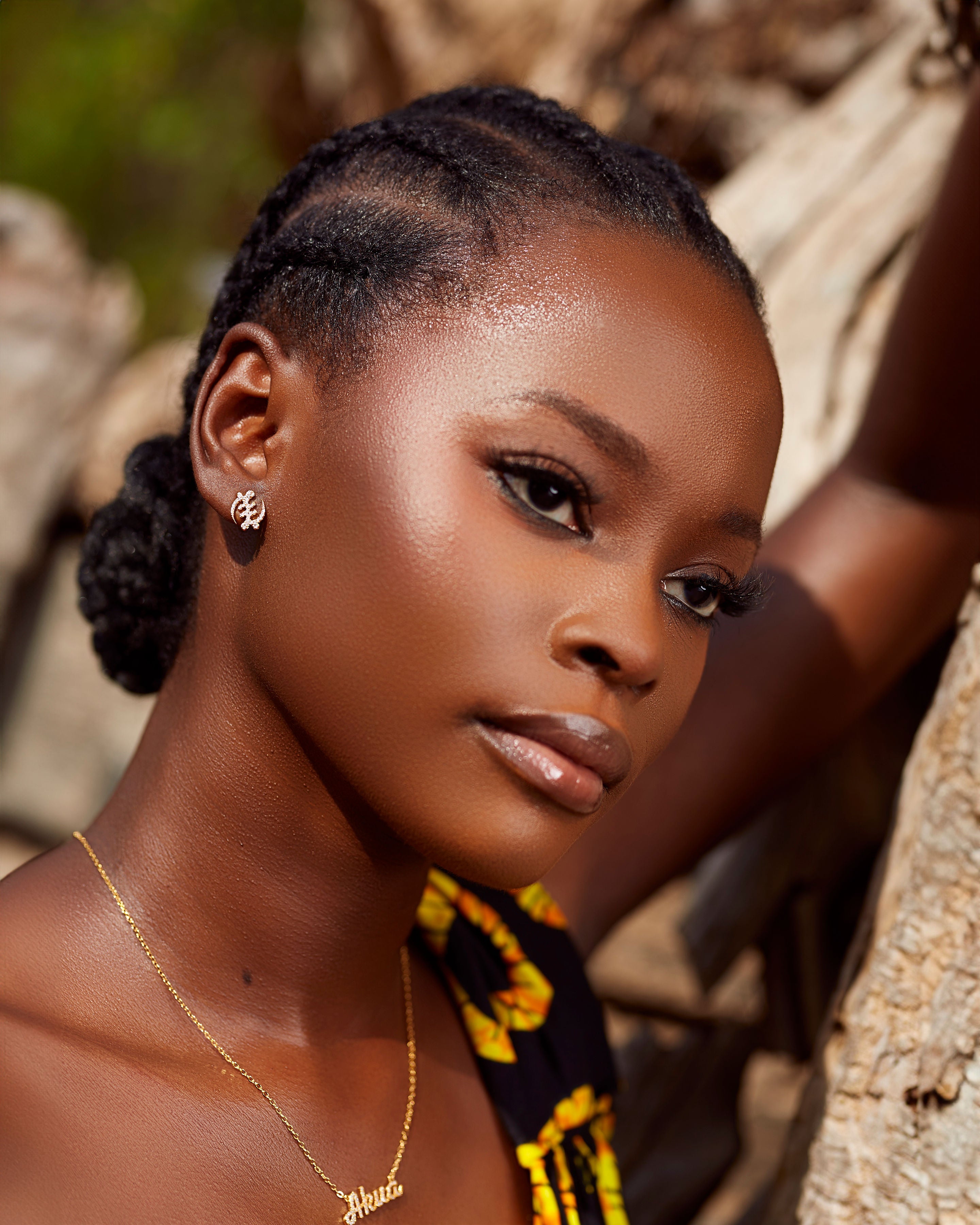 A portrait of a person with braided hair gathered into a bun, wearing CASA London's 18k gold plated Gye Nyame Pavé Earrings Mini. The studs, featuring the iconic Adinkra symbol encrusted with shimmering pavé zircon stones, are complemented by a delicate gold nameplate necklace. The setting is outdoors with natural sunlight highlighting the jewelry's radiance.