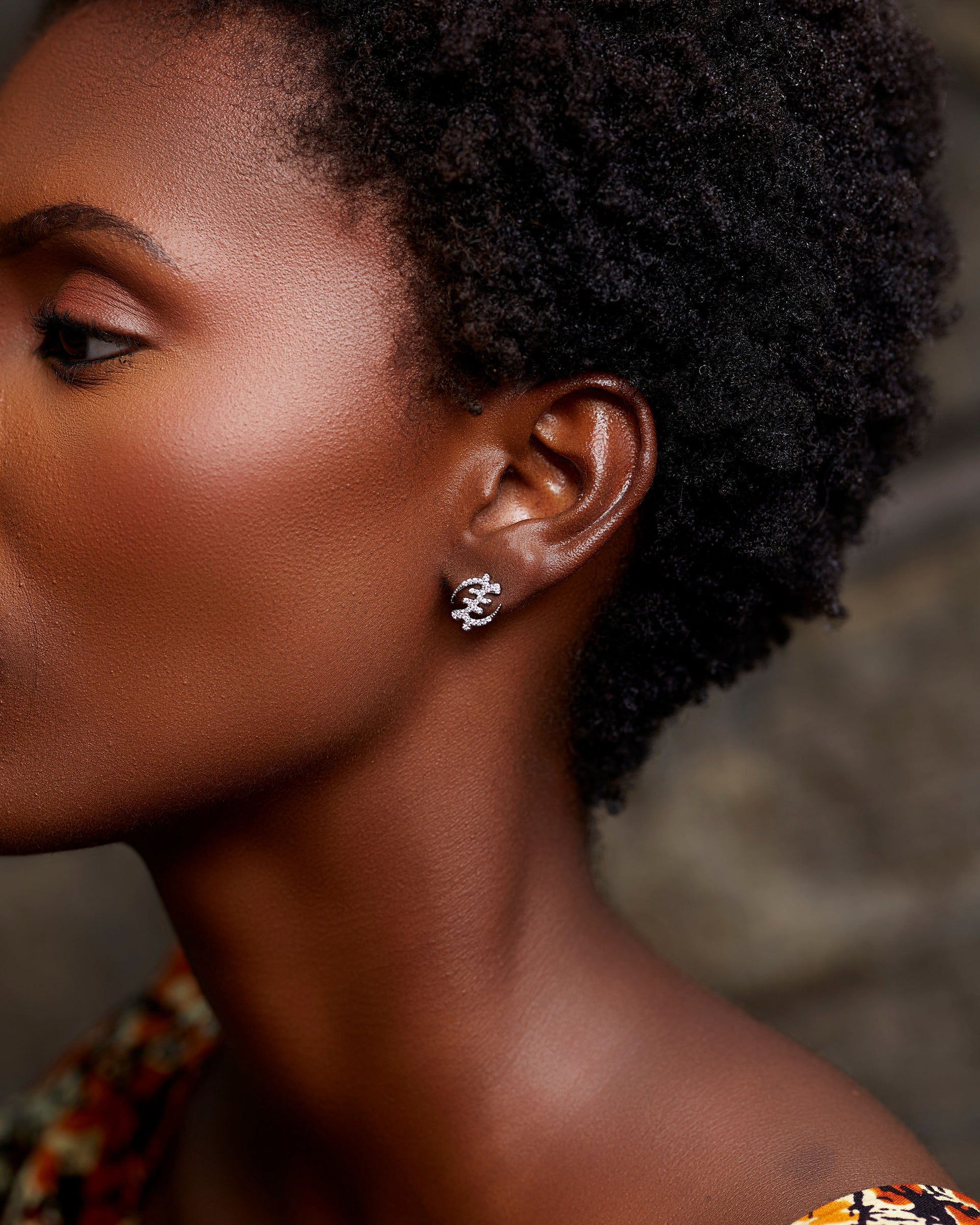 A close-up profile portrait of a person with short, natural dark hair, wearing the Silver Gye Nyame Pavé Earrings Mini. The stud earring, shaped as the iconic Gye Nyame symbol, is encrusted with brilliant pavé zircon stones that sparkle against the soft, warm-toned lighting.