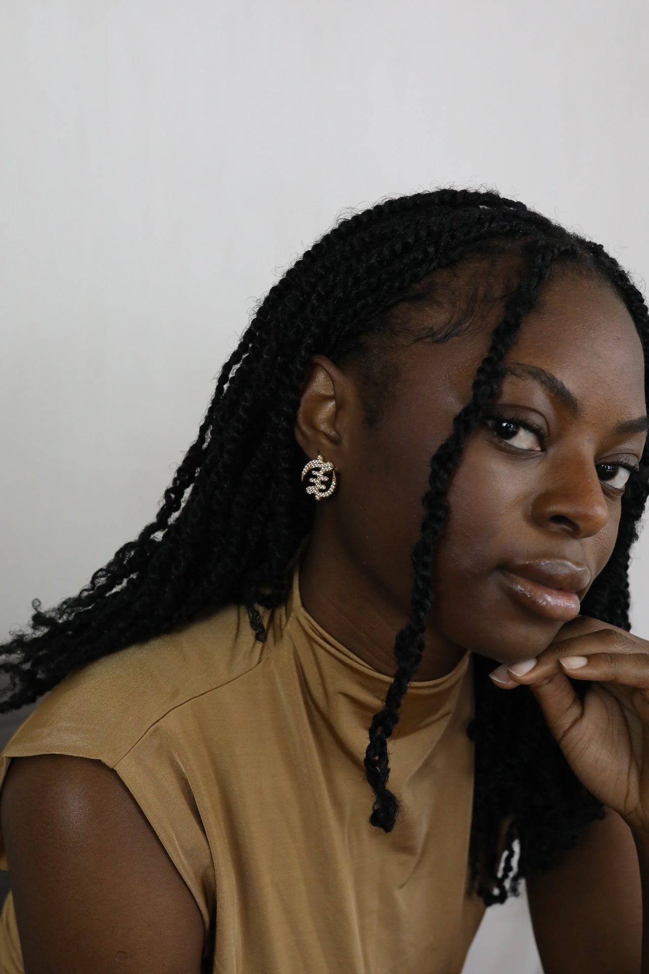 Woman with braided hair wearing gold Gye Nyame Adinkra symbol stud earrings with pavé-set high-grade zircon stones. Model is looking from the side.