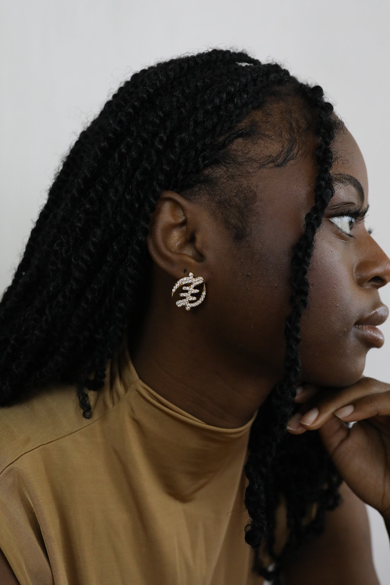 Woman with braided hair wearing gold Gye Nyame Adinkra symbol stud earrings with pavé-set high-grade zircon stones. Model is looking from the side.