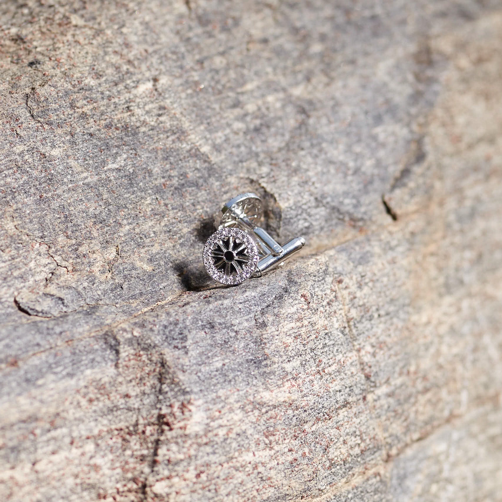 A detailed shot of CASA London's Ohene Men's Pavé Black Enamel Cufflinks resting on the textured surface of a grey rock. The circular cufflinks feature a striking black enamel center with radiating silver spokes, surrounded by a border of shimmering pavé-set stones. The natural outdoor lighting highlights the intricate design and the brand's commitment to merging cultural heritage with sophisticated, modern luxury.