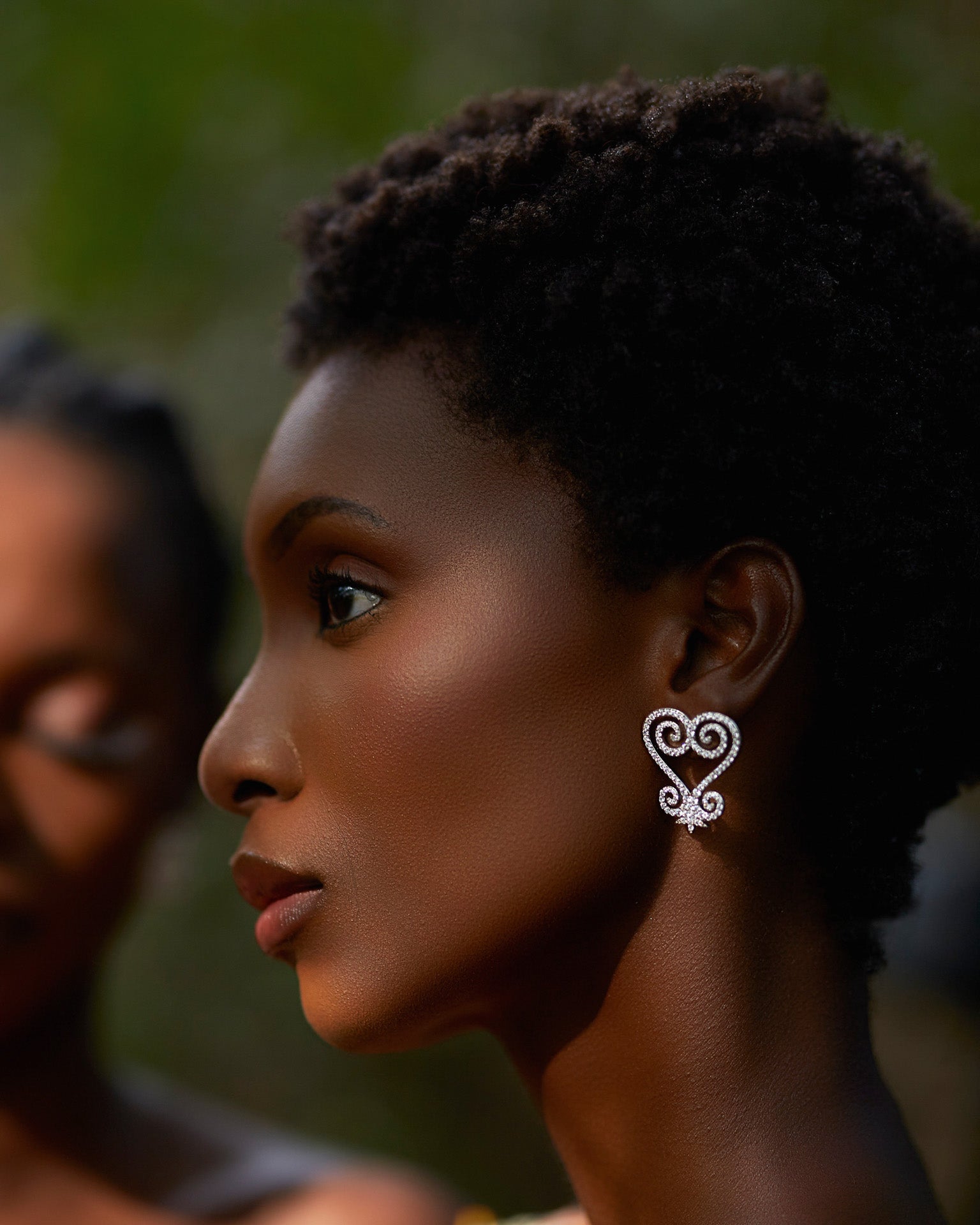A close-up profile of a model wearing CASA London's Sankofa Pavé Silver earrings. The earrings feature the intricate, heart-shaped Sankofa Adinkra symbol, meticulously encrusted with shimmering pavé-set stones that catch the soft, natural light. The model’s natural hair and the soft-focus background highlight the brand's commitment to blending cultural heritage with contemporary luxury.