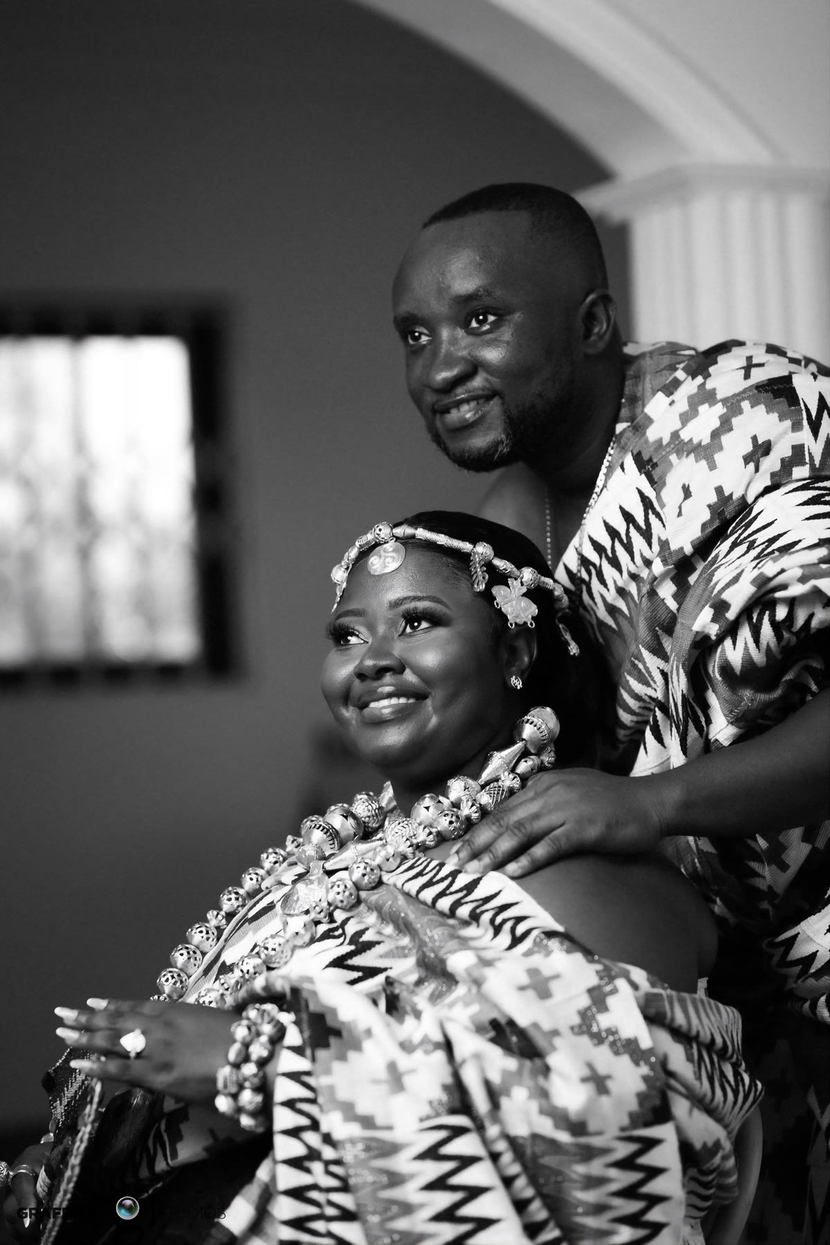 Black and white portrait of a bride in a beaded headband and layered beaded necklaces, her husband in kente behind her with a hand on her shoulder