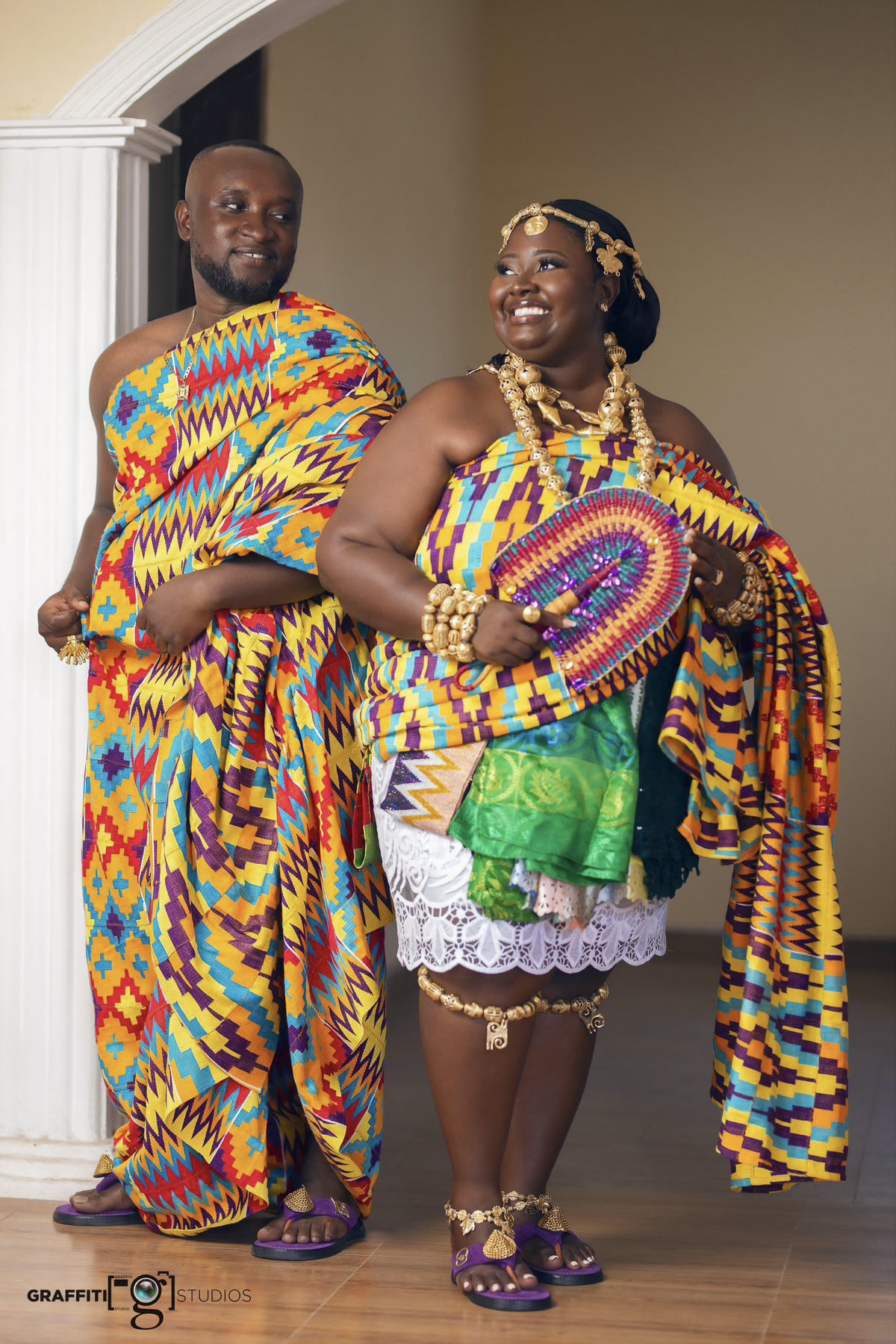 A couple standing in full-length kente, she holding a patterned fan and both wearing gold at the ankle, neck and wrist