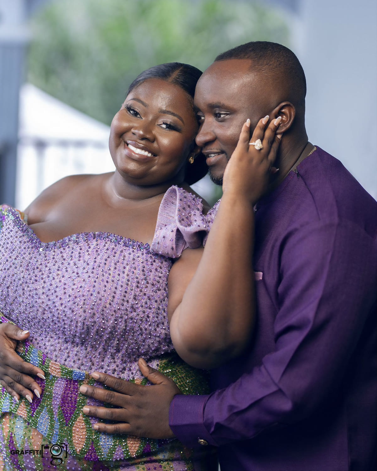 A couple embracing, she in a lilac gown and he in a purple shirt with his hand on her cheek, a ring on her finger