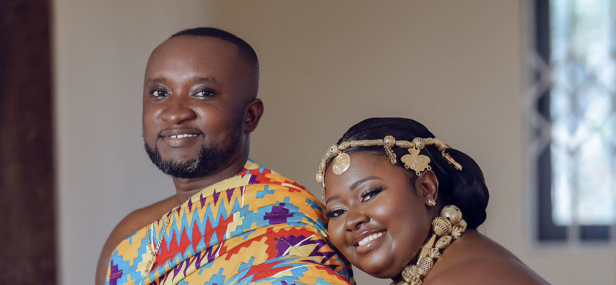 A Ghanaian couple in bright kente, the groom smiling and the bride leaning against him wearing a gold Adinkra headpiece and a beaded gold necklace